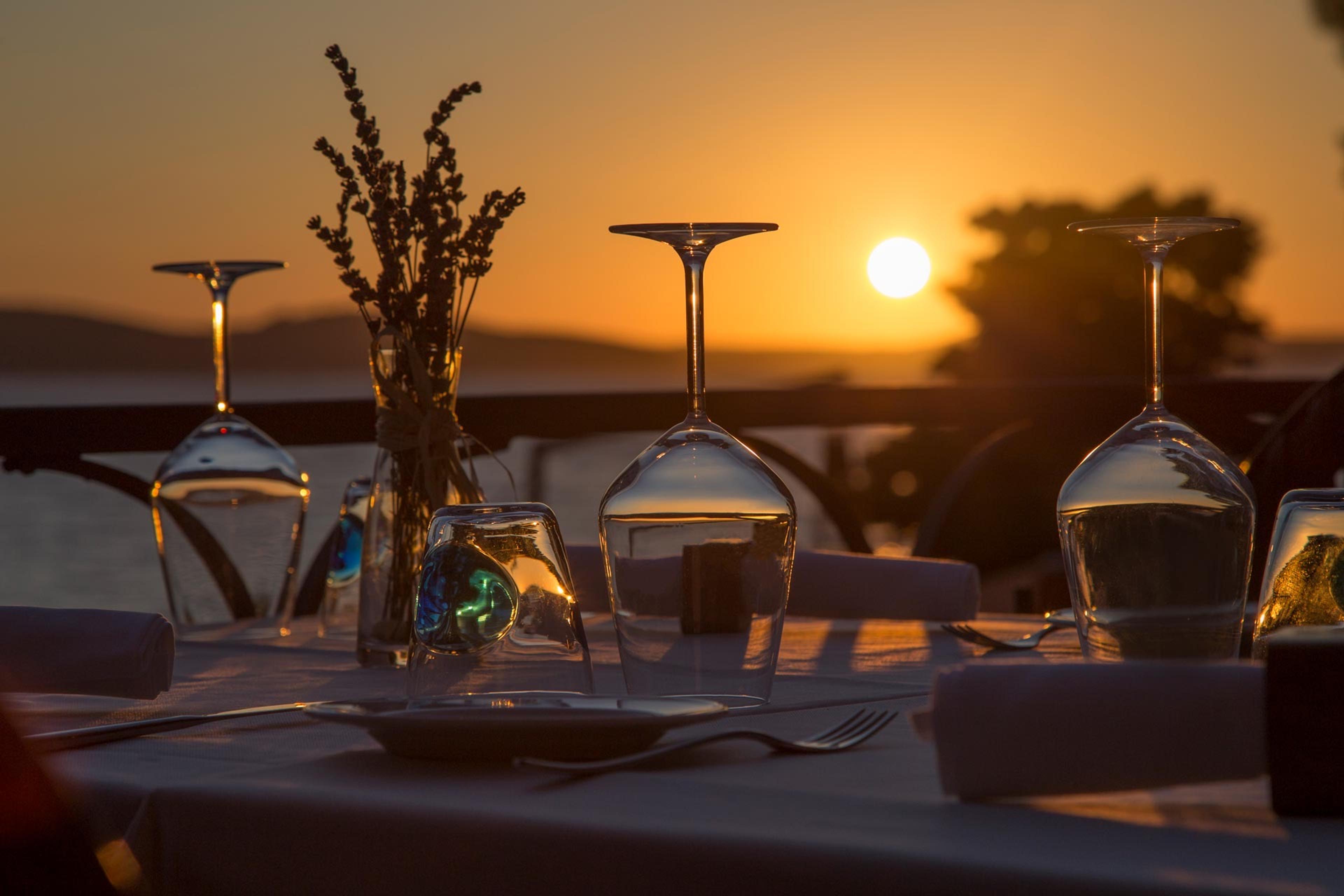 Wine glasses and a table set for dinner on a terrace, with the sun setting low over water and hills in the background.