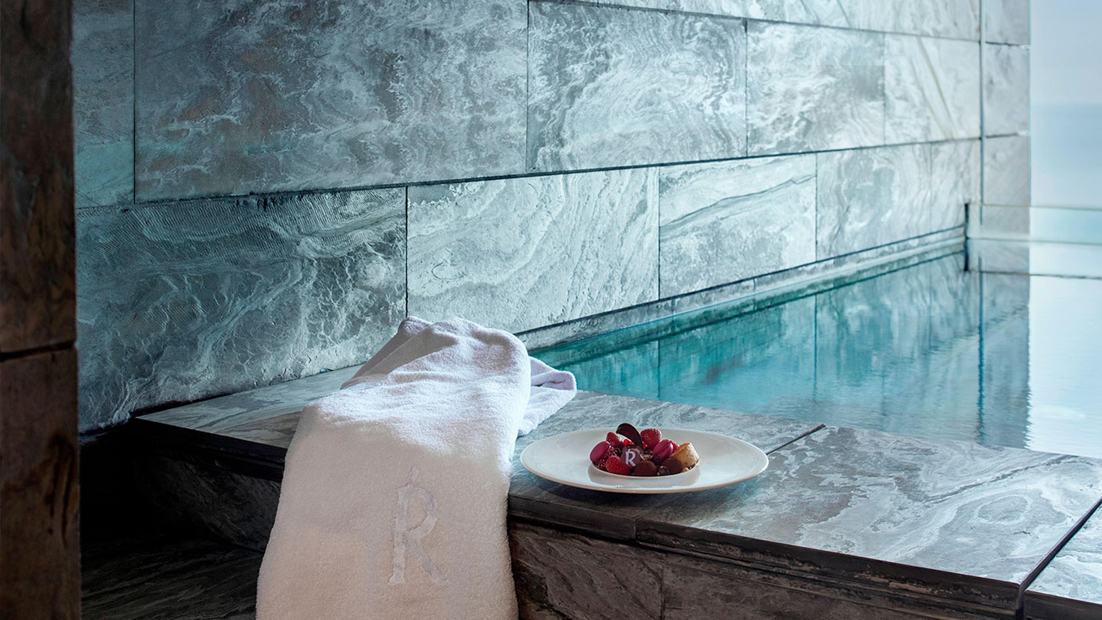 The tiled edge of an infinity pool with a folded monogrammed towel and a plate of berries and pastry on the stone surround, the sea beyond.