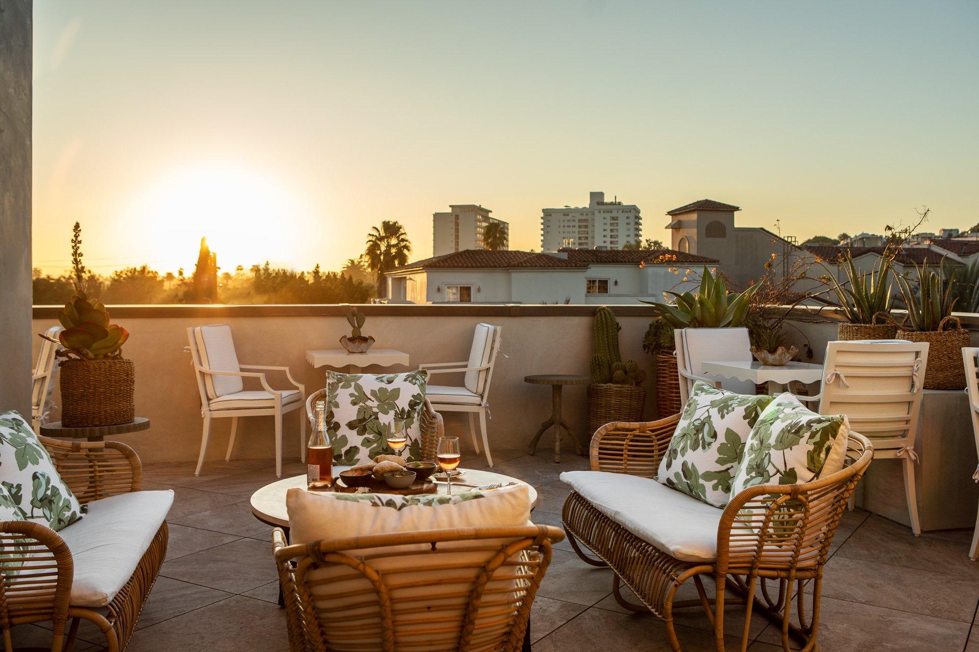 A rooftop terrace with wicker chairs and a low table set with wine and snacks, overlooking the city skyline at sunset.