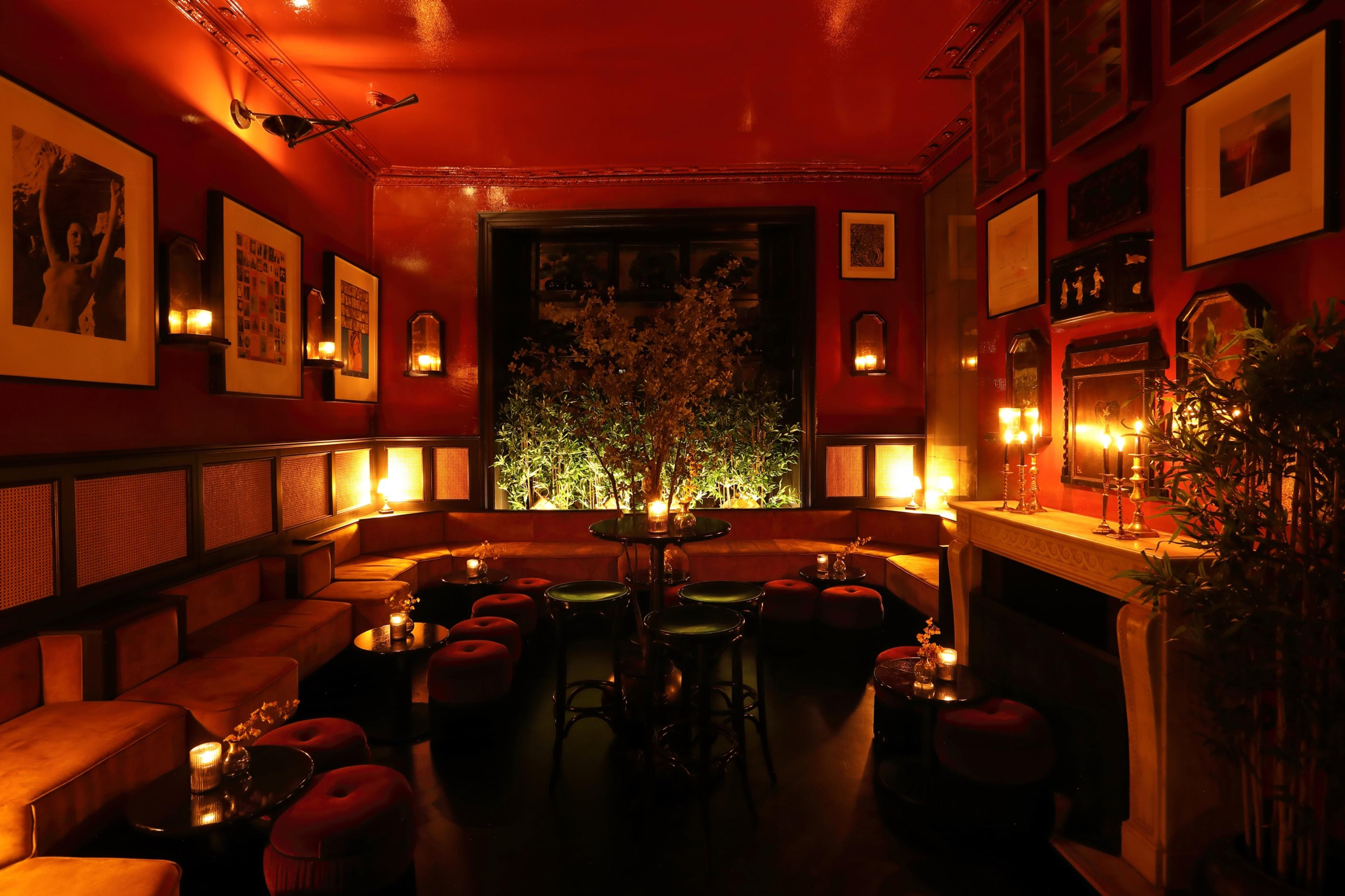 A red-walled lounge with low velvet sofas, round stools, candlelit side tables and a lit fireplace, with a small tree visible through an arched window.