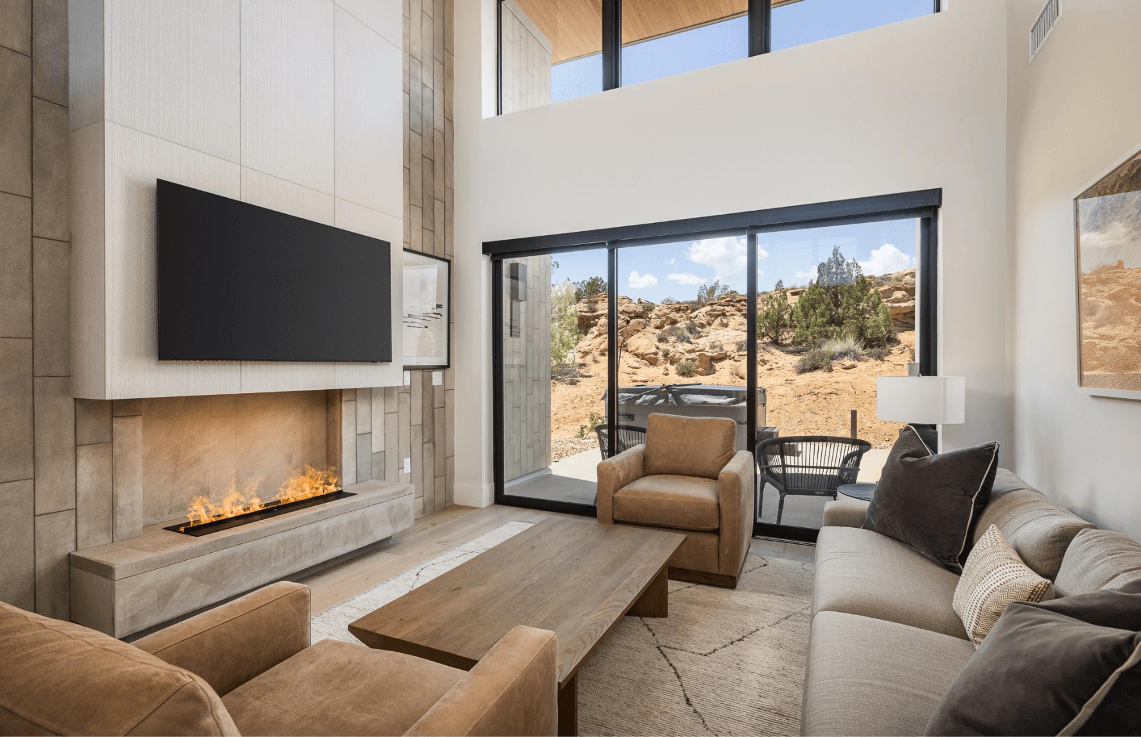 A living room with a lit fireplace below a wall-mounted television, leather armchairs, a sofa, and glass doors open to a patio and hot tub with red rock hills beyond.