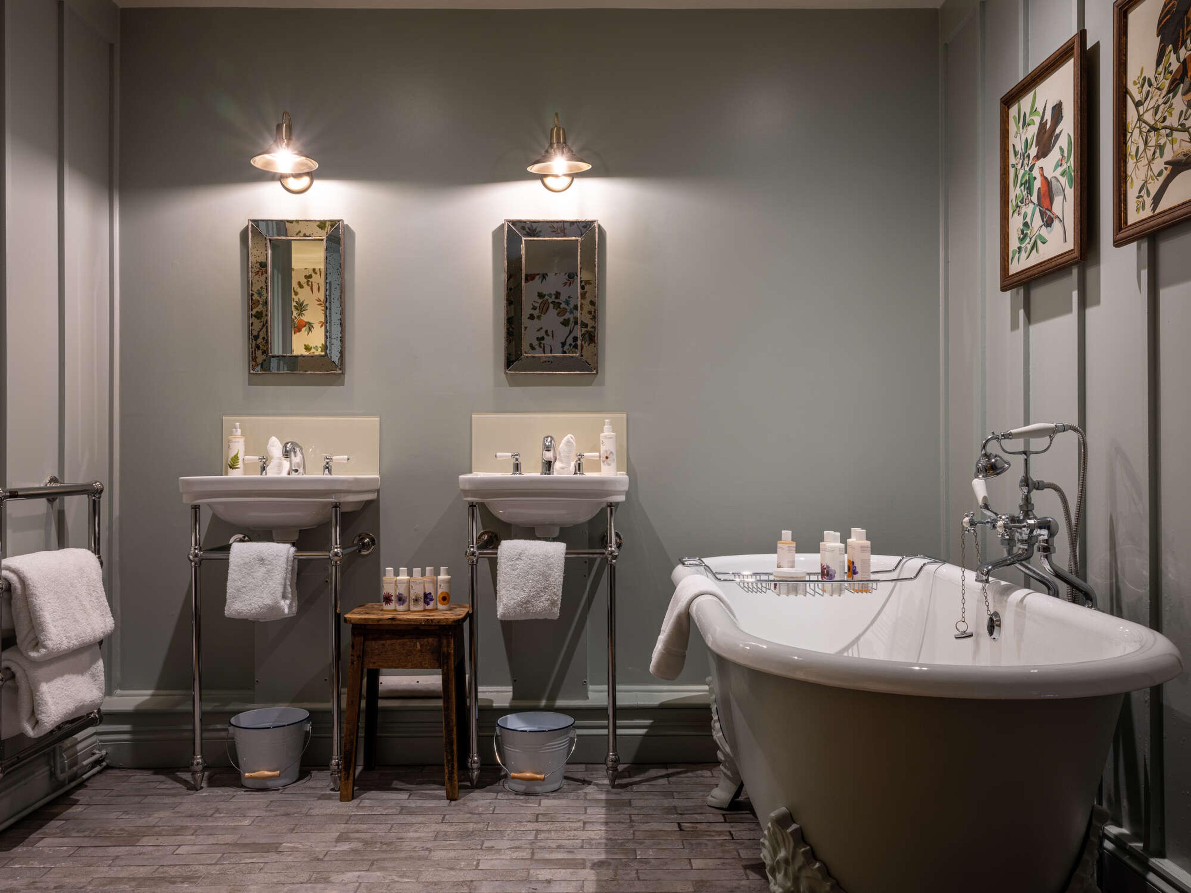A bathroom with two pedestal sinks under lit wall mirrors, a roll-top tub, framed bird prints and towels on a heated rail.