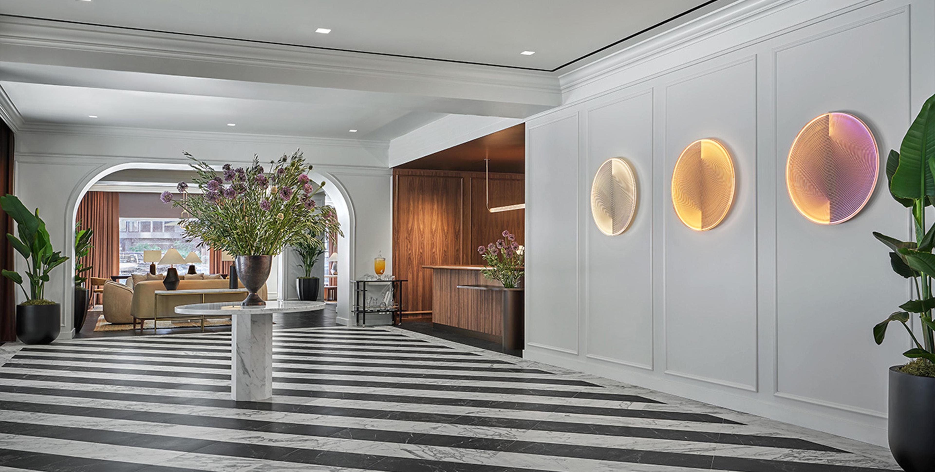 A hotel lobby with black and white striped marble floors, a round table with a large flower arrangement, and arched doorways leading to a bar.