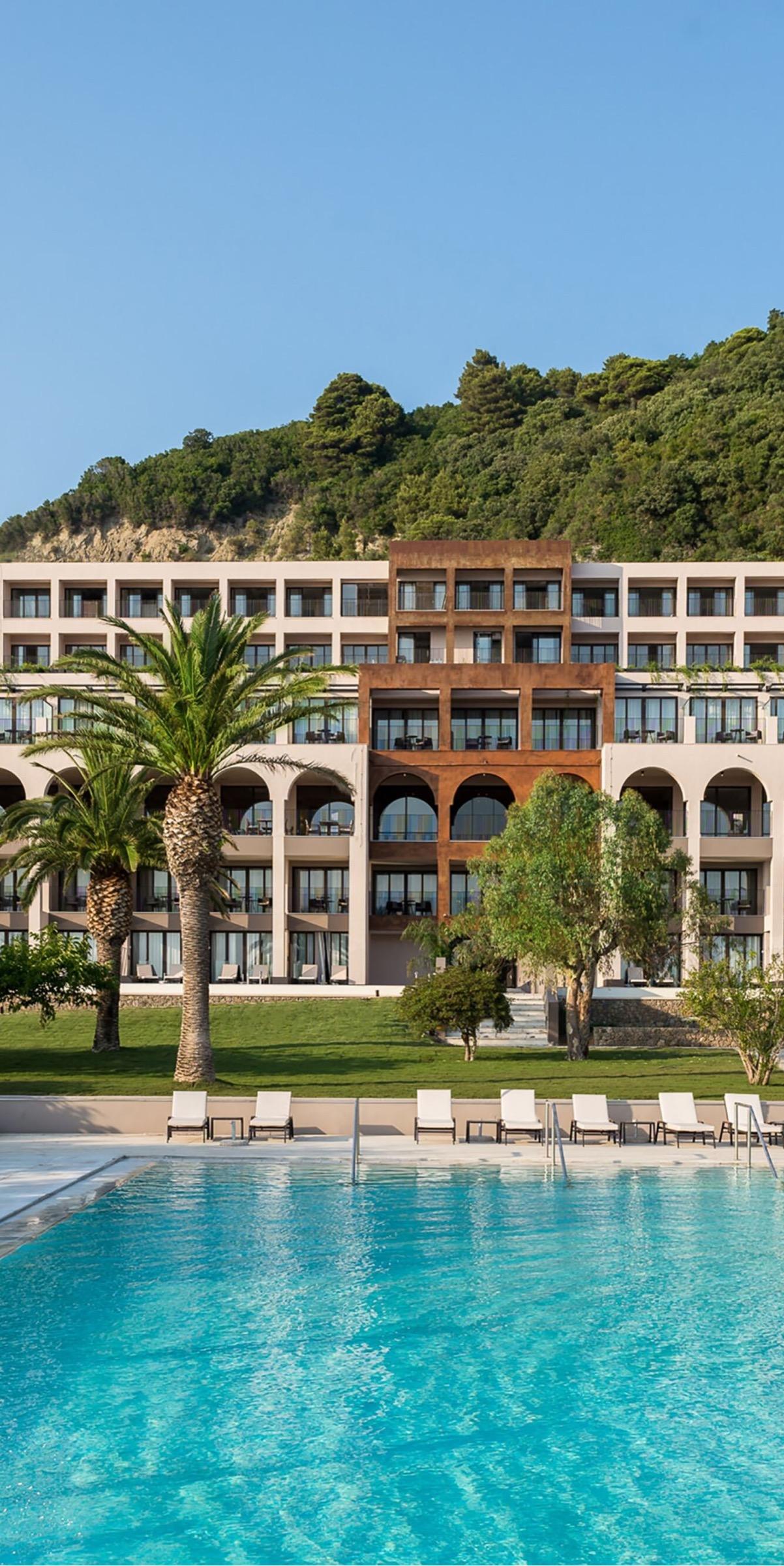 The hotel's cream and rust-colored facade seen full width across a lawn, with two palm trees and a row of white sunbeds beside the pool.