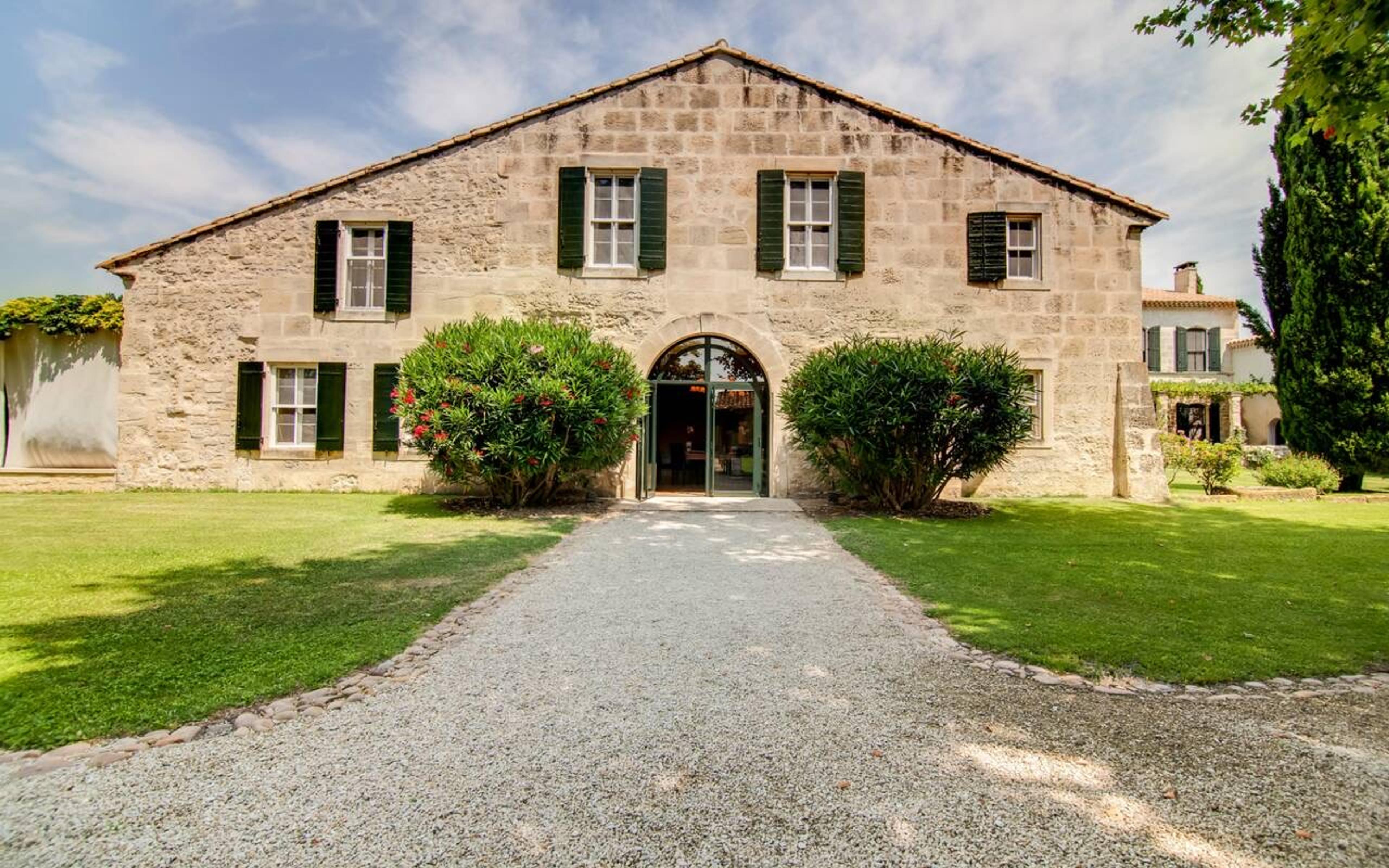 A stone farmhouse with green shutters and an arched doorway, seen across a lawn with a gravel path leading to the entrance.