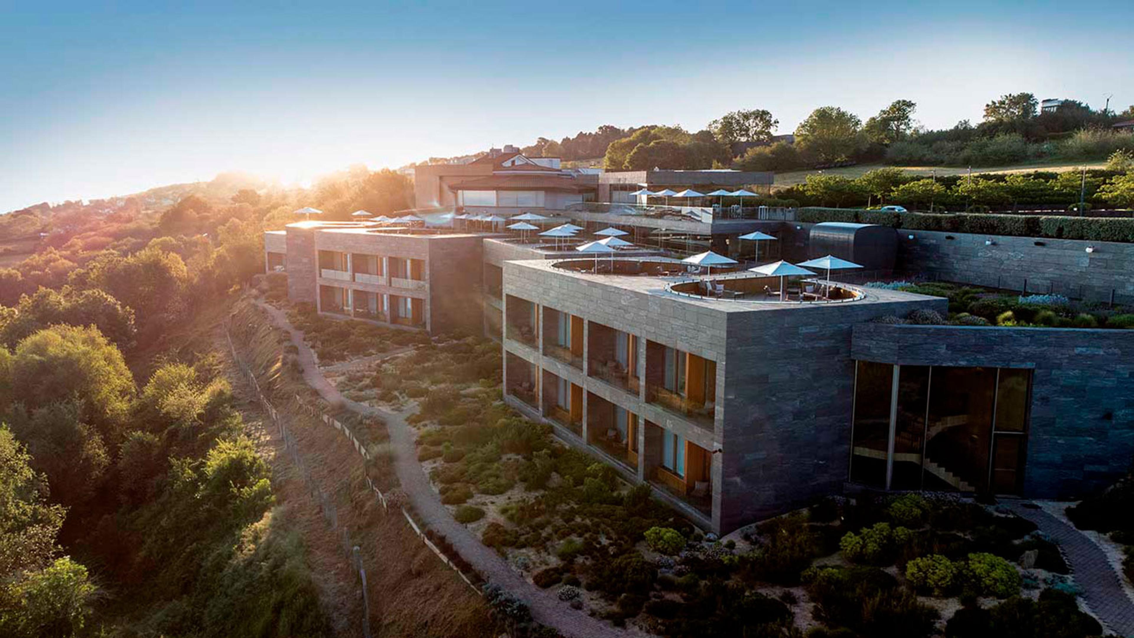 An aerial view of the hotel's stone wings and rooftop pool terrace at golden hour, sun flaring over wooded hills, with gardens below.