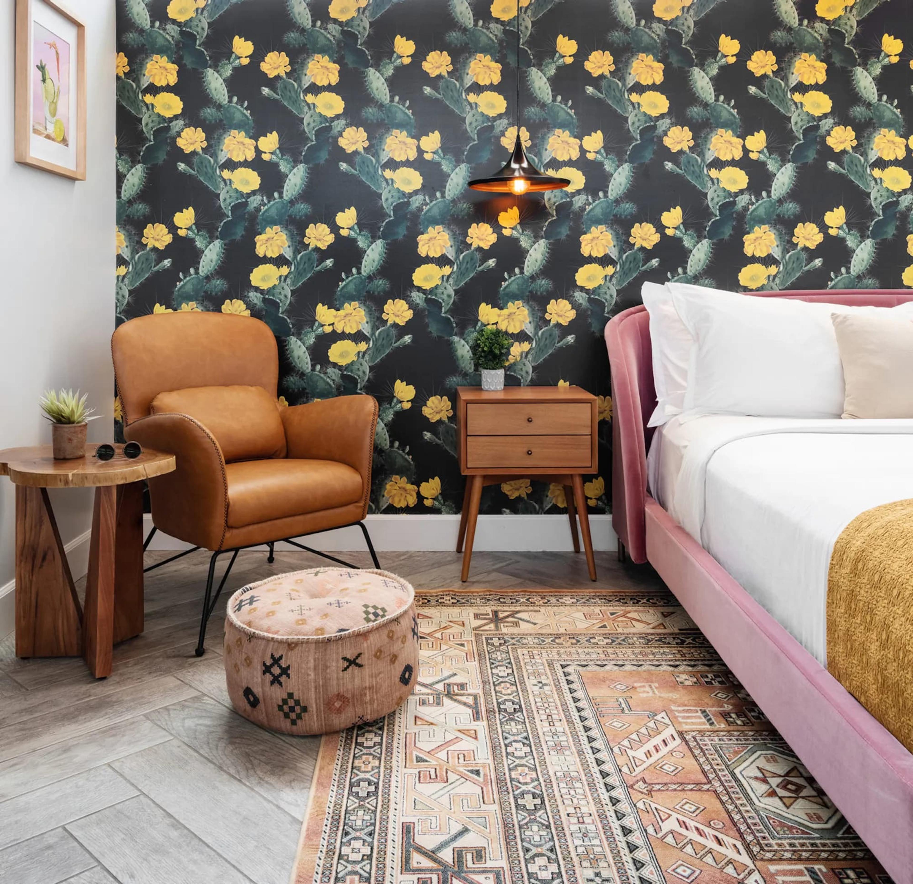 A corner of a hotel room with a tan leather armchair, a round wooden side table with a small succulent on it, and a patterned rug on a grey herringbone floor, against black wallpaper printed with yellow cactus flowers.