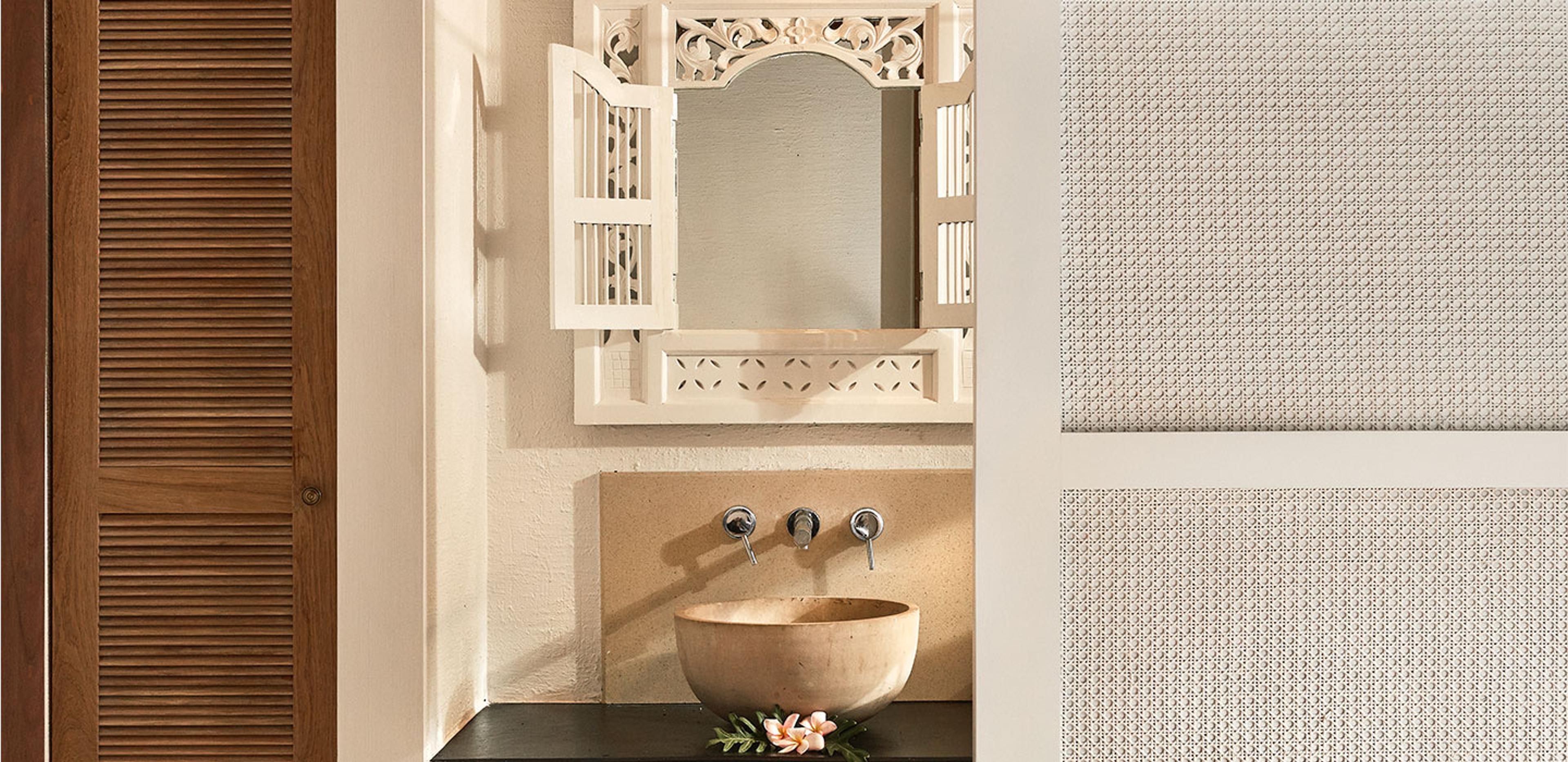 A carved wooden mirror hangs above a round stone basin set into a dark wood vanity, with a frangipani flower resting on the ledge.