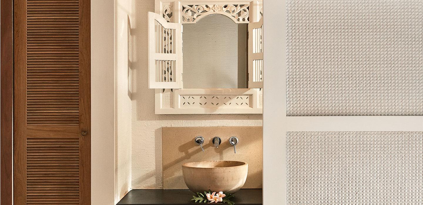 A carved wooden mirror hangs above a round stone basin set into a dark wood vanity, with a frangipani flower resting on the ledge.