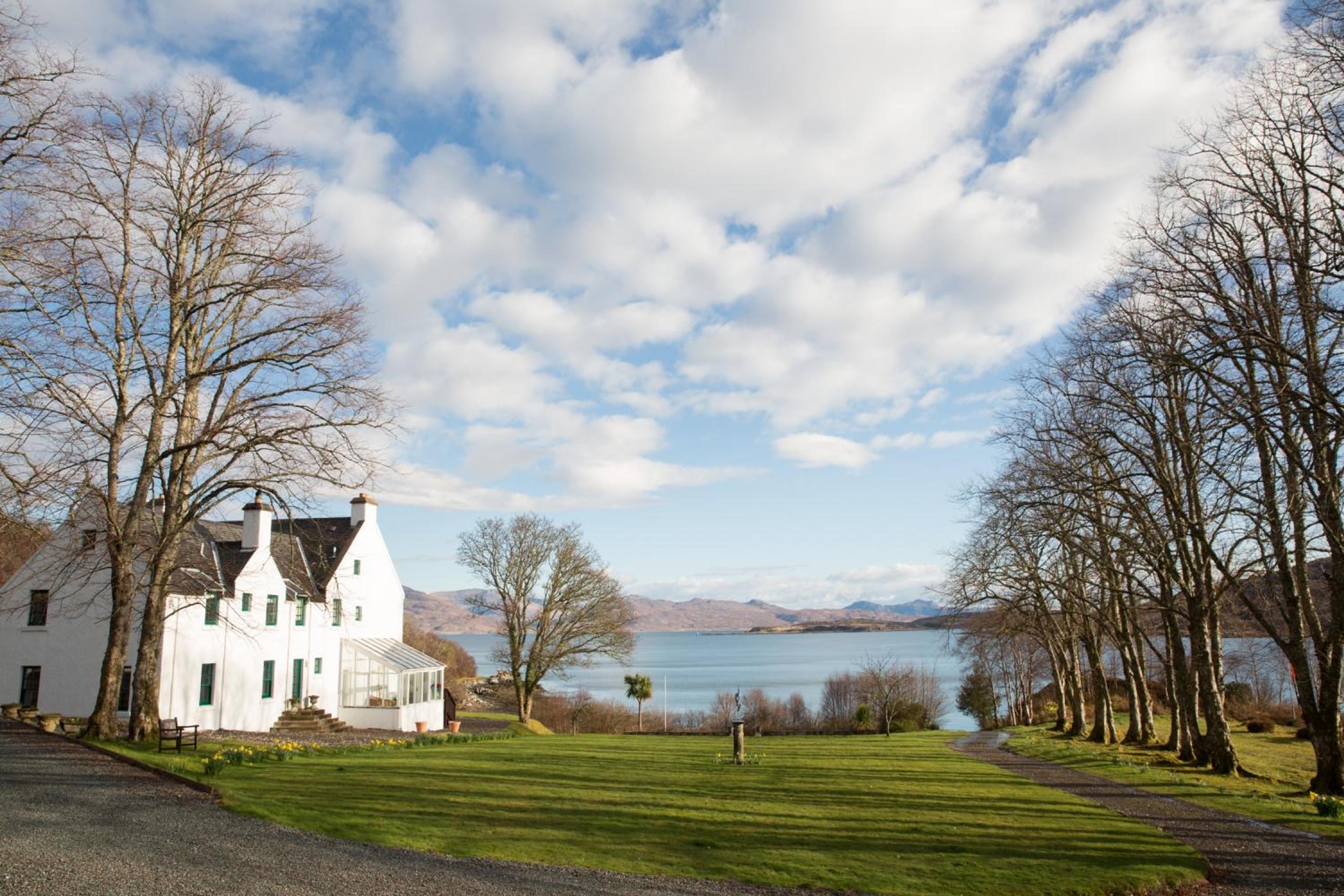 A white harled house beside a sea loch under a wide sky of scattered cloud, framed by bare trees on a lawn running down to the water.