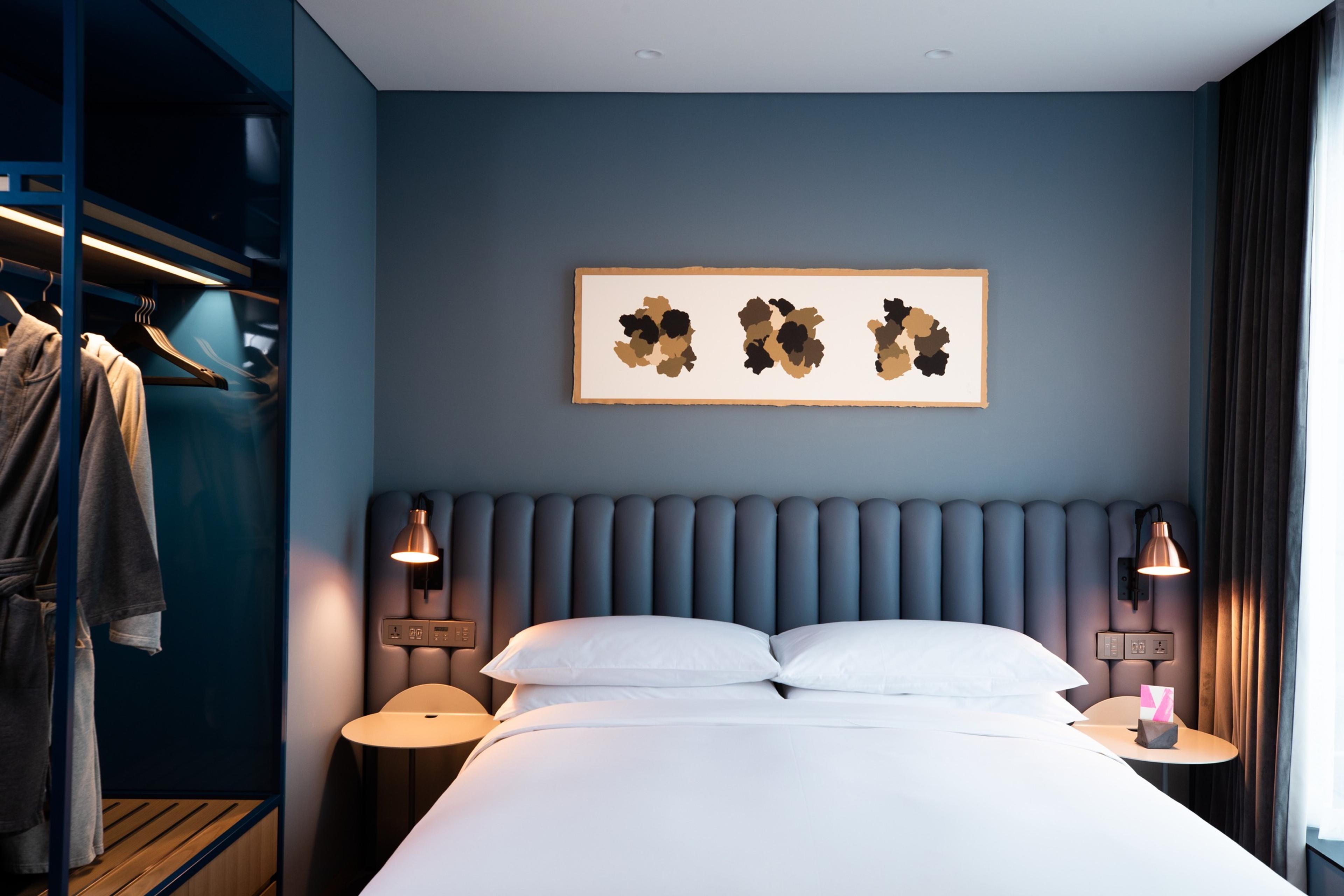 A hotel bedroom with a channel-tufted blue headboard, a bed made up in white linen and an open closet with a robe hanging beside it.