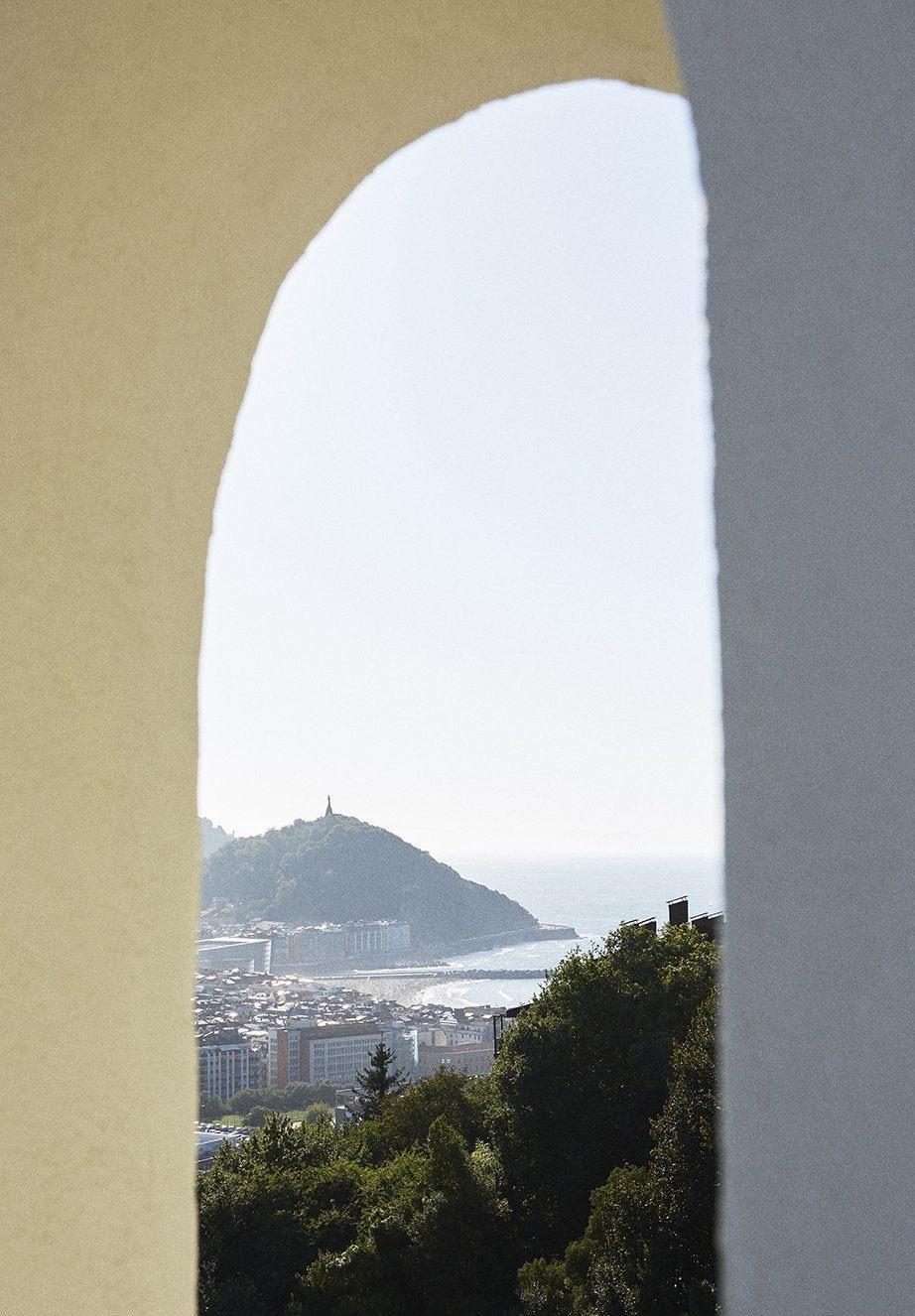 a wooded hill topped with a statue, seen through a white stone archway with the sea and town below
