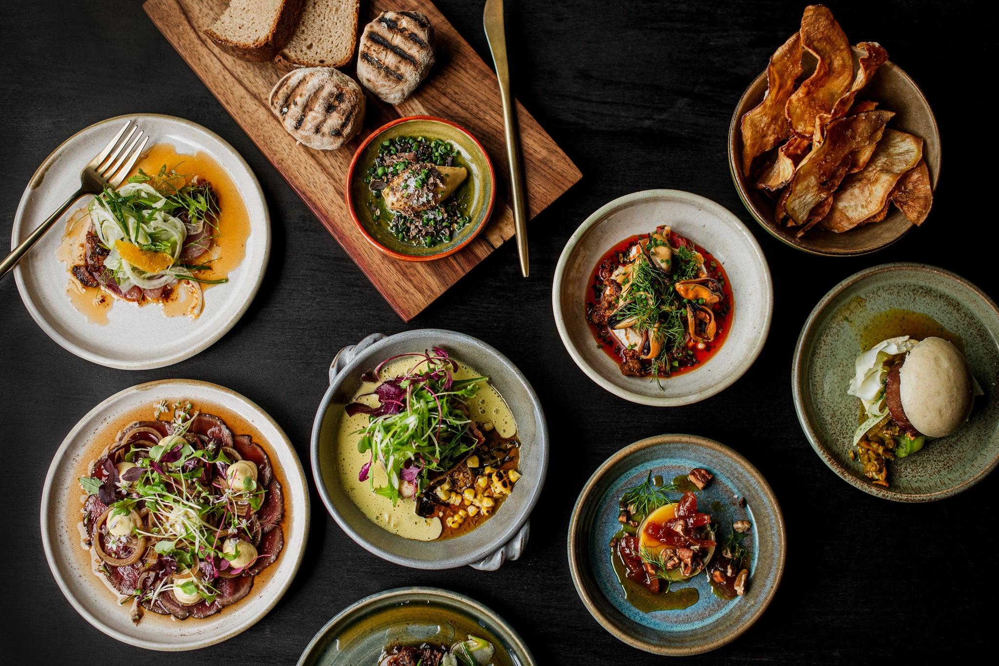 Small plates on a dark table, seen from above: cured meat, bread on a board and a bowl of crisps