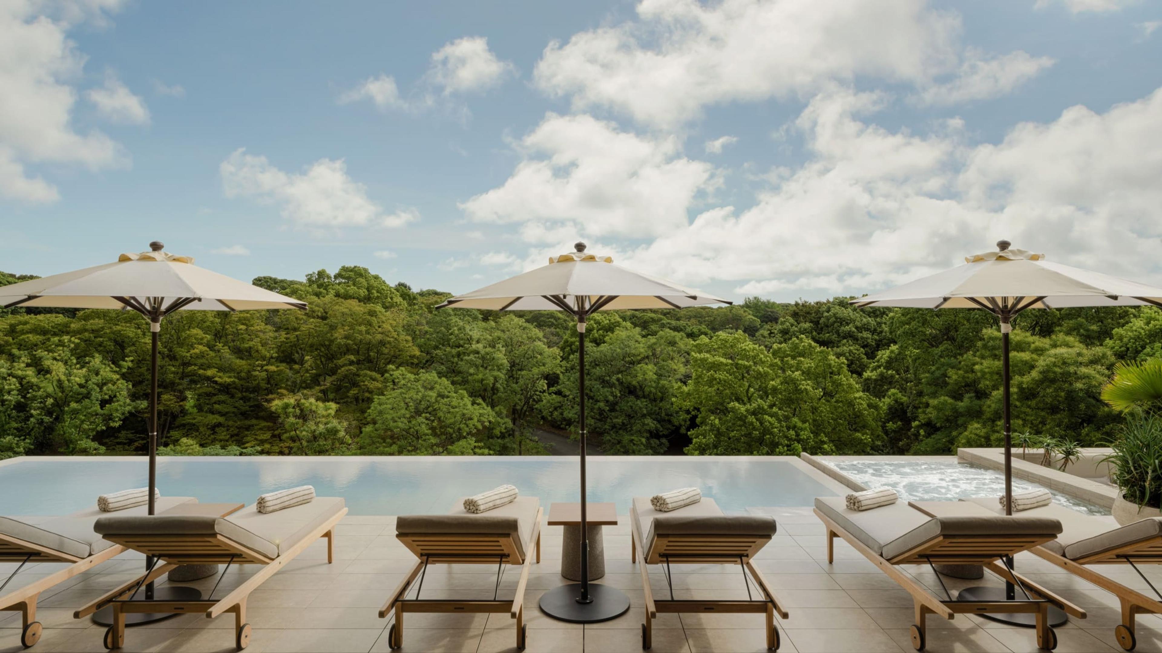 a rooftop infinity pool lined with cushioned loungers and cream umbrellas, backed by a dense tree canopy under a blue sky with clouds