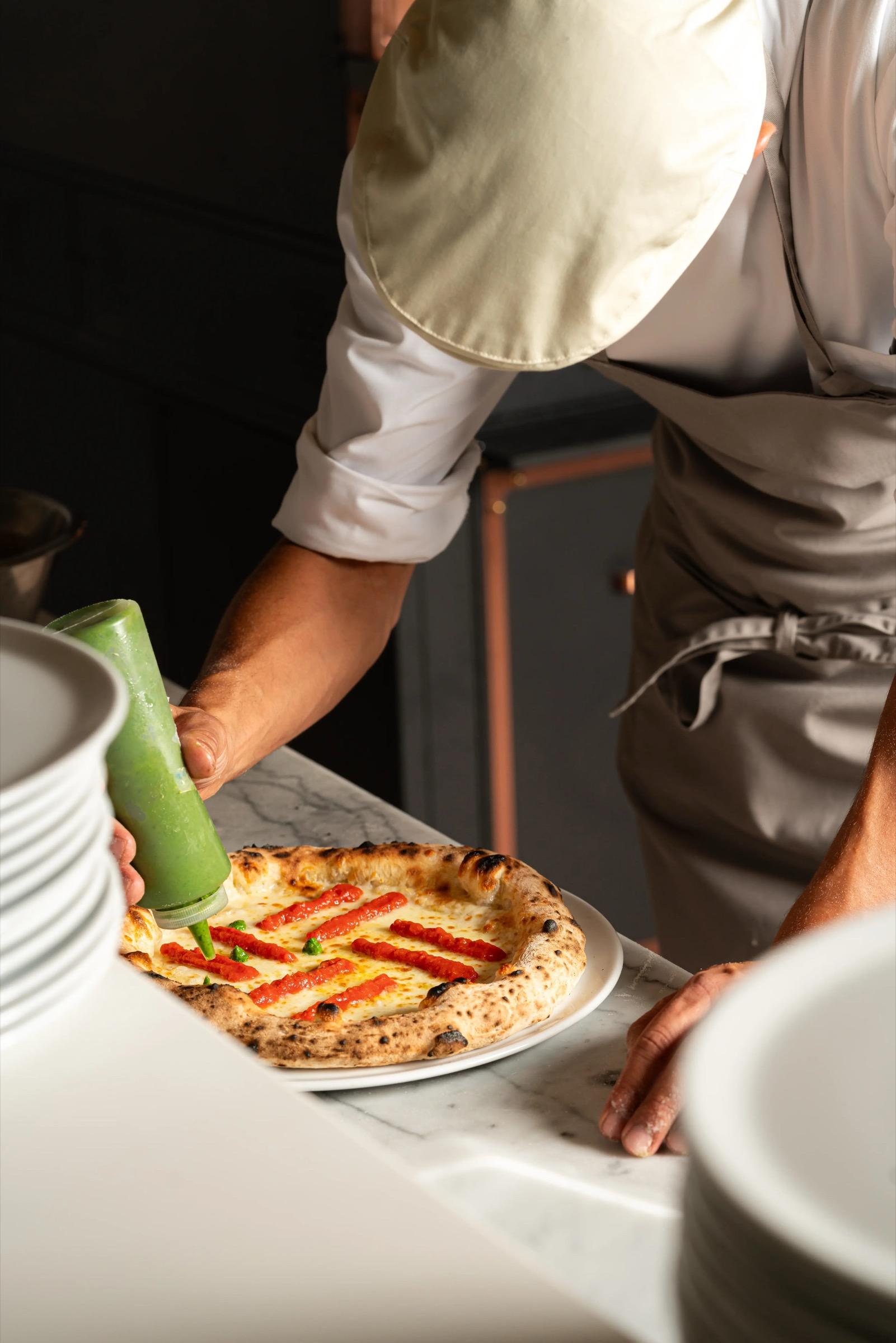 A chef's hands pipe a green sauce onto a wood fired pizza on a marble counter.