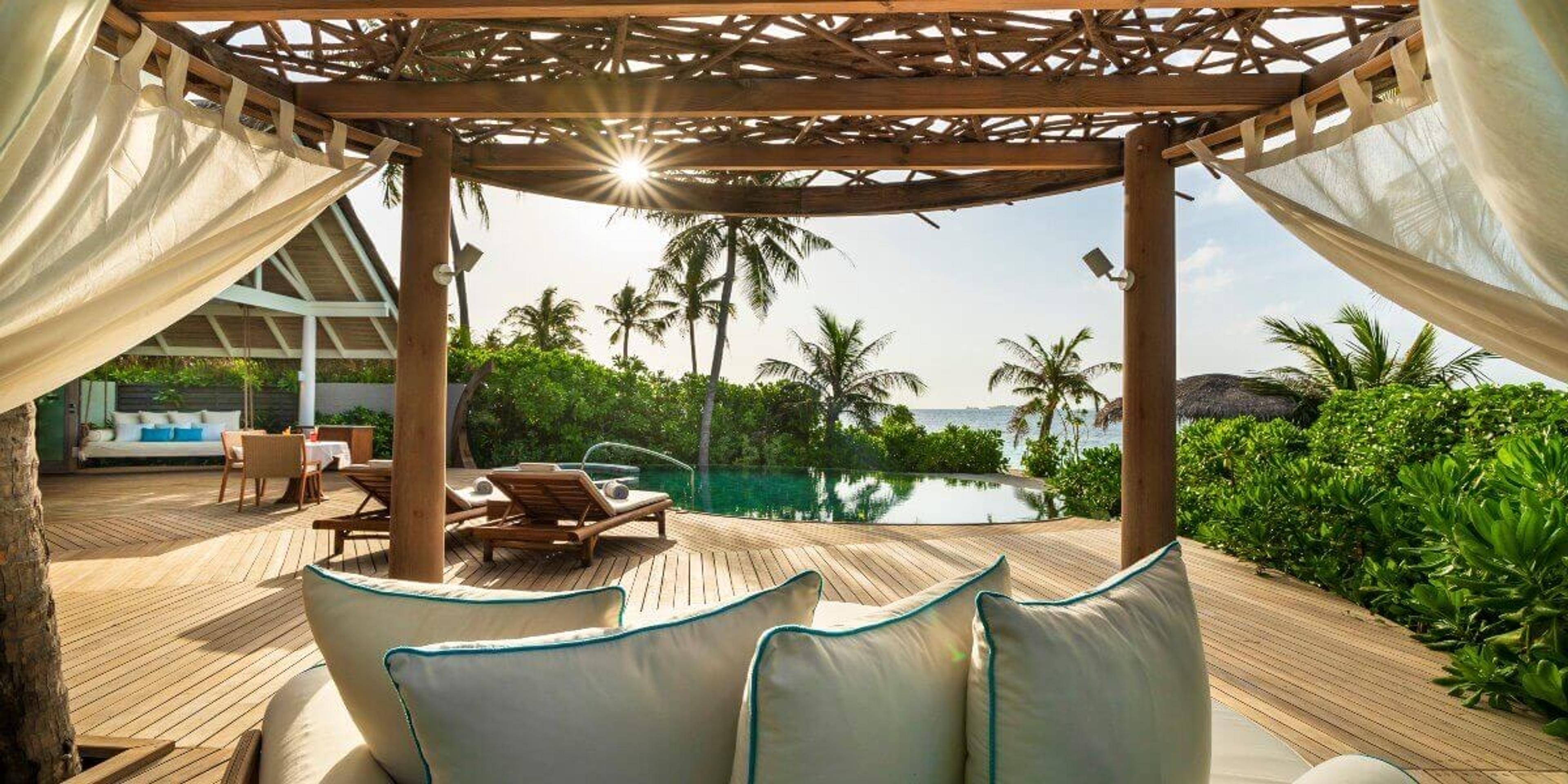 A shaded daybed under a woven canopy beside an infinity pool, palm trees and the sea lit by low sun.