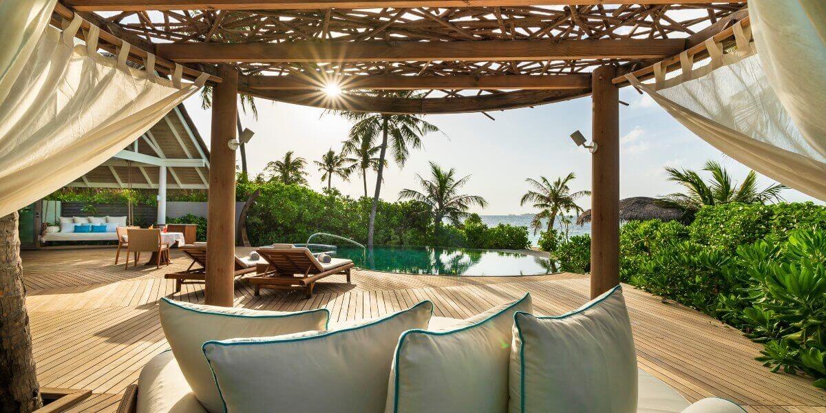 A shaded daybed under a woven canopy beside an infinity pool, palm trees and the sea lit by low sun.