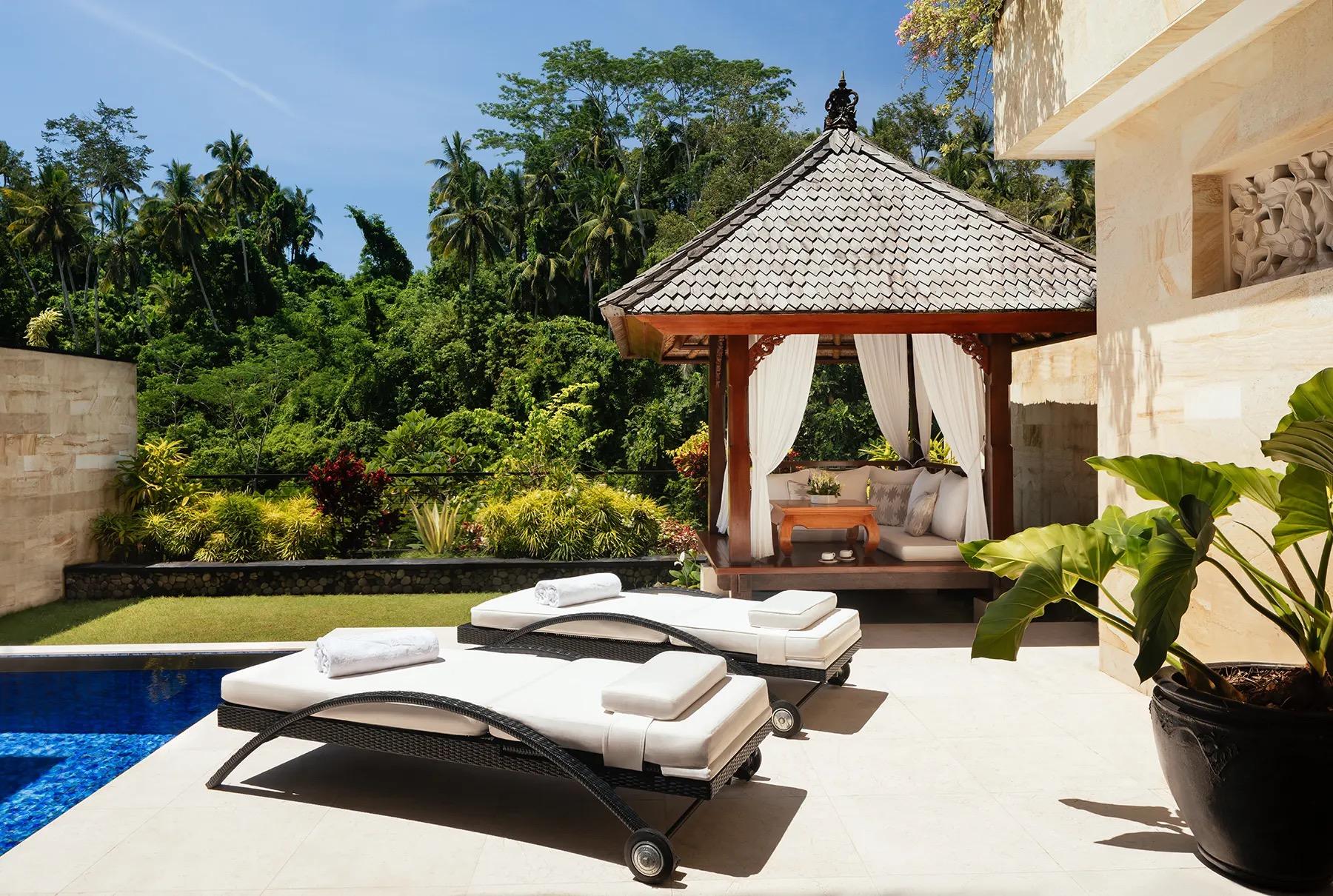 Sunloungers and a stone gazebo with a statue sit on a terrace overlooking a forested valley.