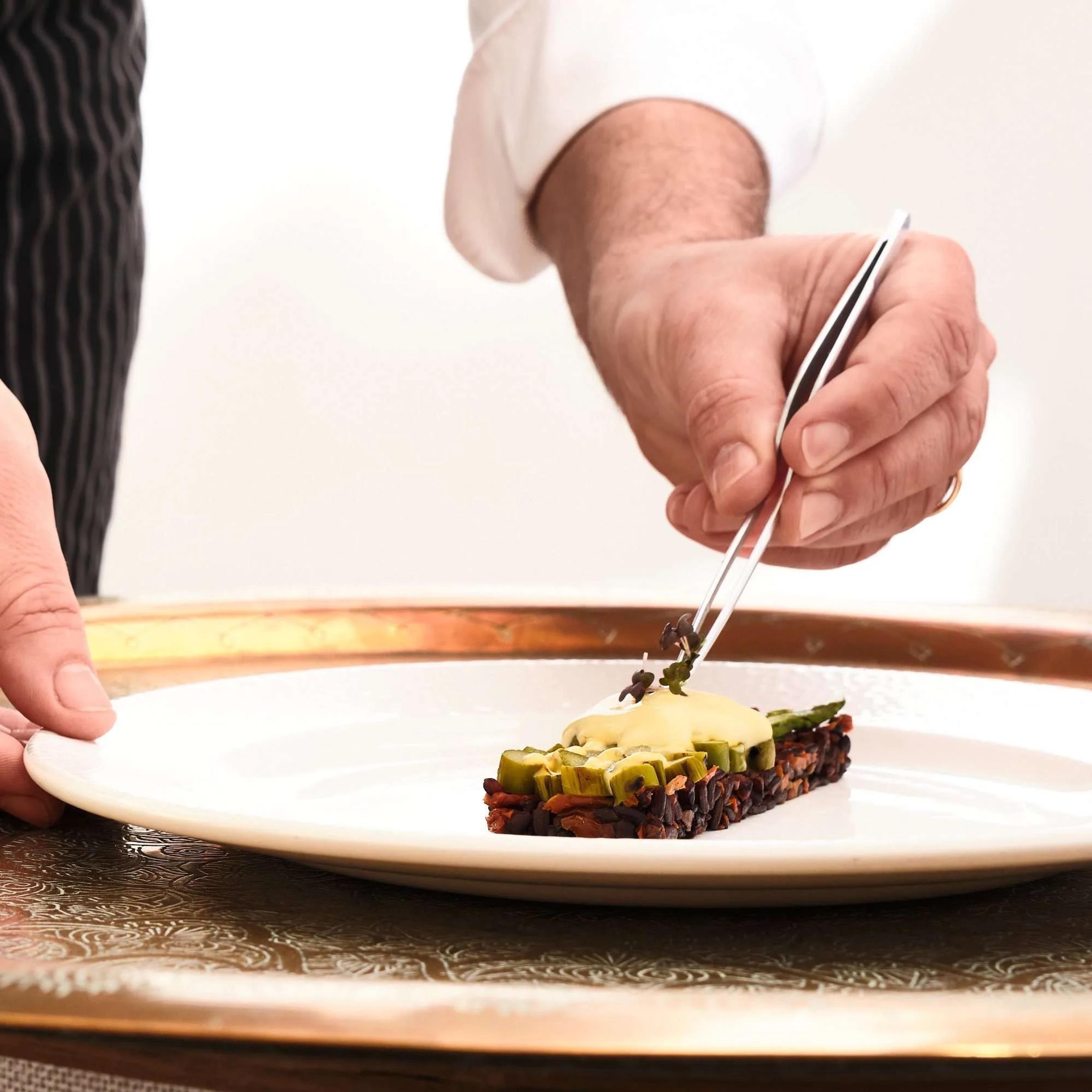 A chef's hand placing a garnish with tweezers on a slice of fish on a white plate.