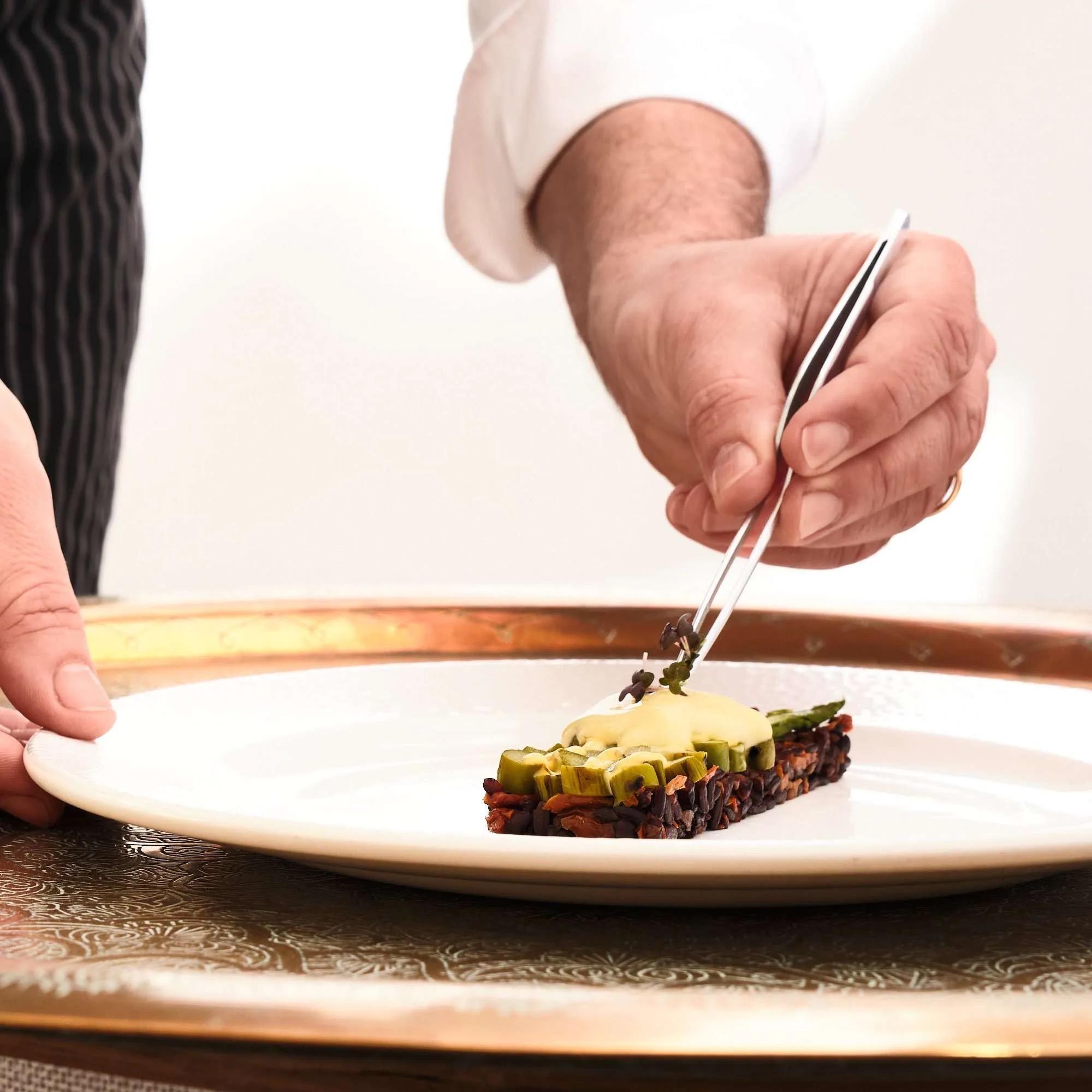 A chef's hand placing a garnish with tweezers on a slice of fish on a white plate.