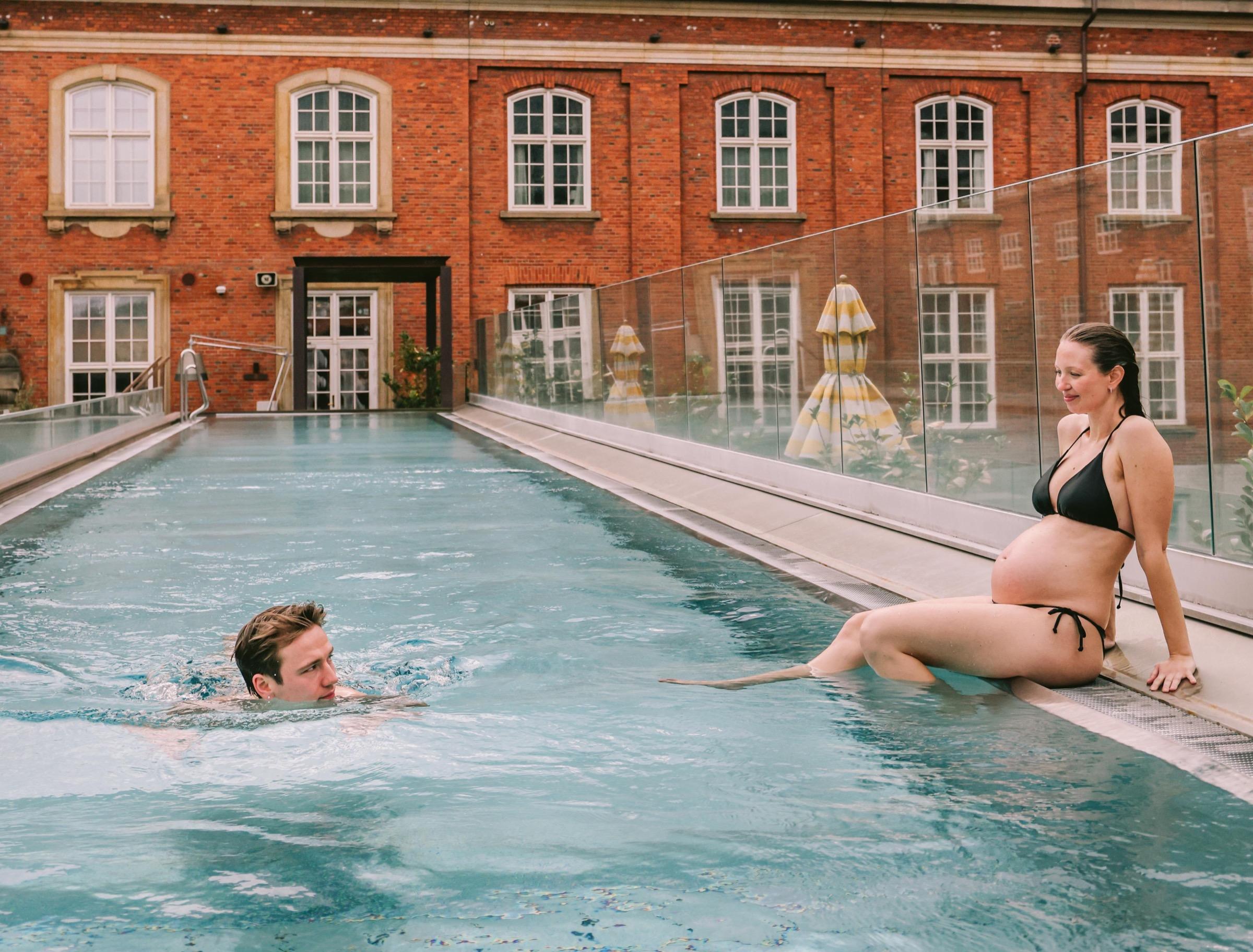 A man swimming and a pregnant woman sitting at the edge of an outdoor pool in front of a red brick building with arched windows.