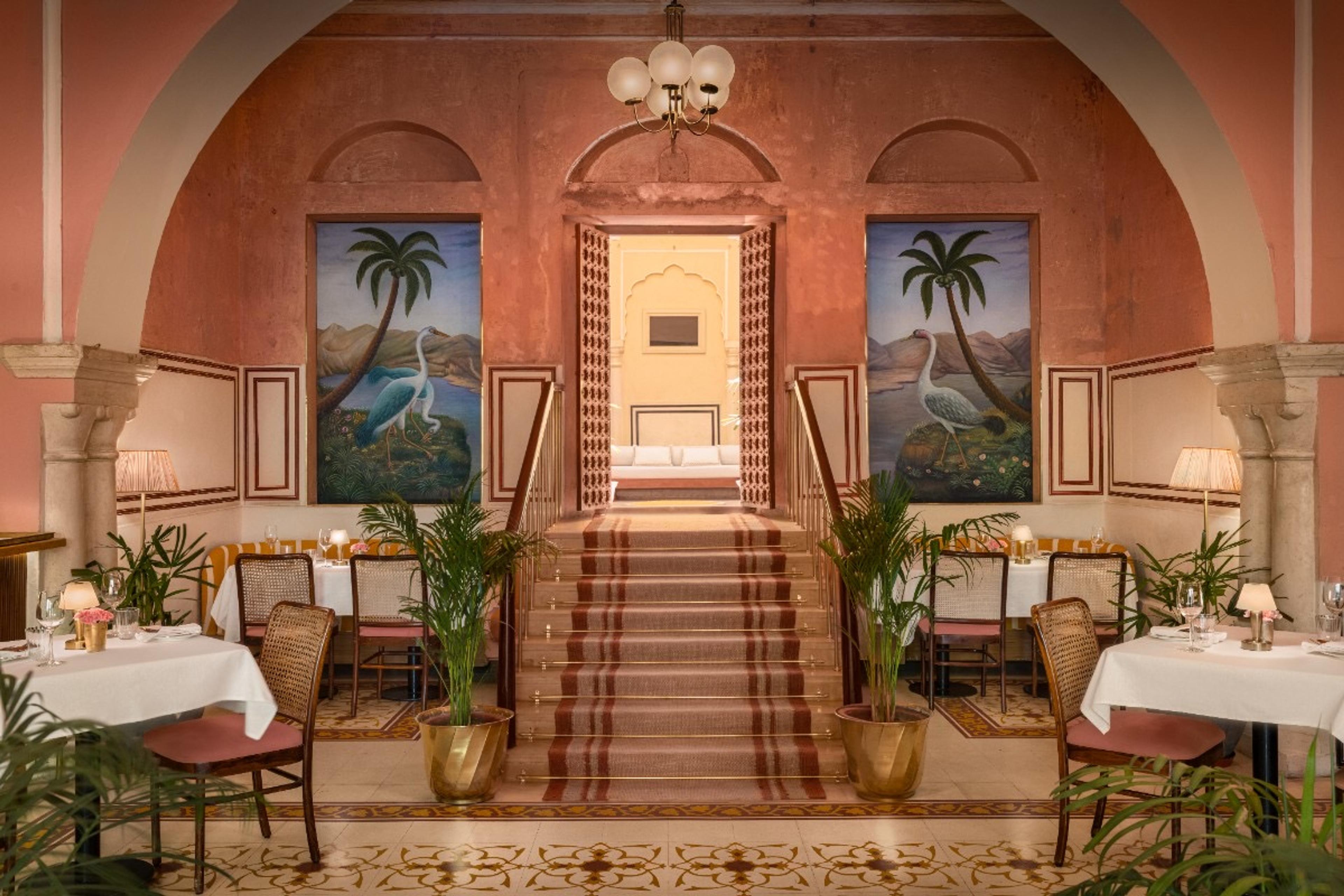 A dining room in terracotta pink with arched doorways, palm paintings and a few laid tables