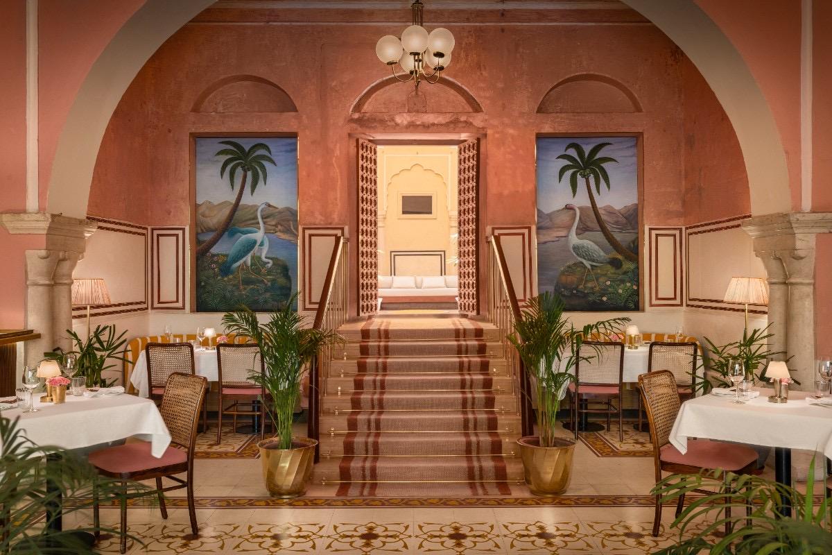A dining room in terracotta pink with arched doorways, palm paintings and a few laid tables