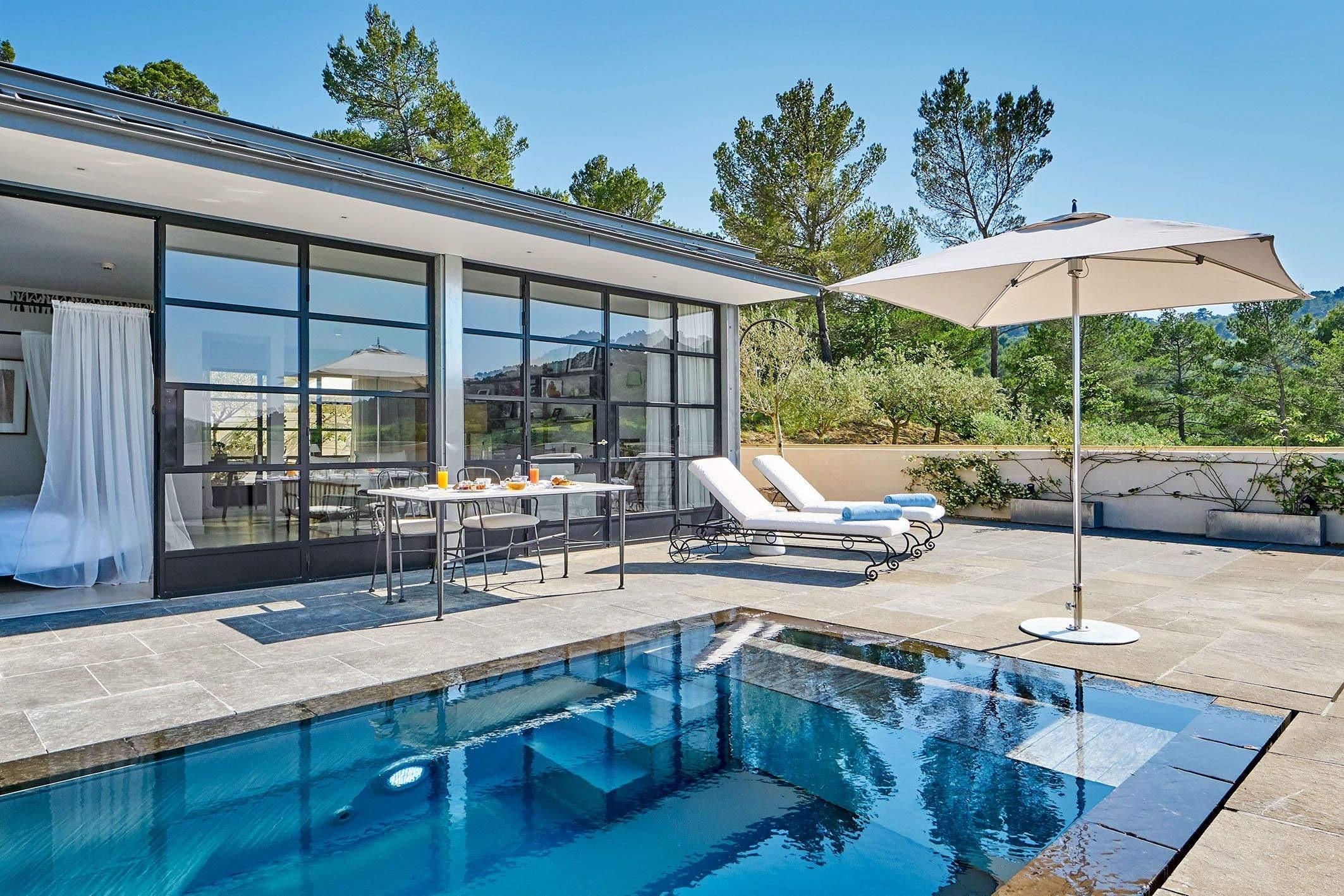 A pool beside a glass-fronted villa, with white loungers and a cream umbrella on the stone terrace.