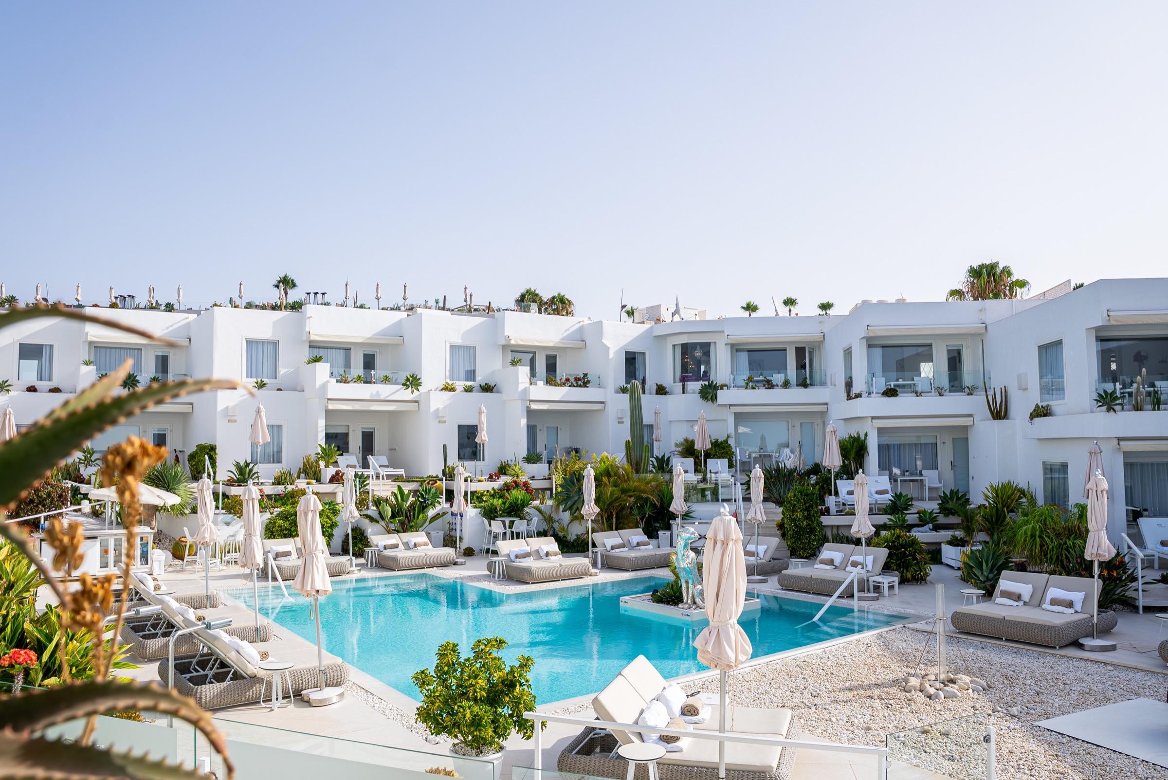 A white multi-level hotel building wrapped around a turquoise swimming pool, with sun loungers, umbrellas and cacti around the water.