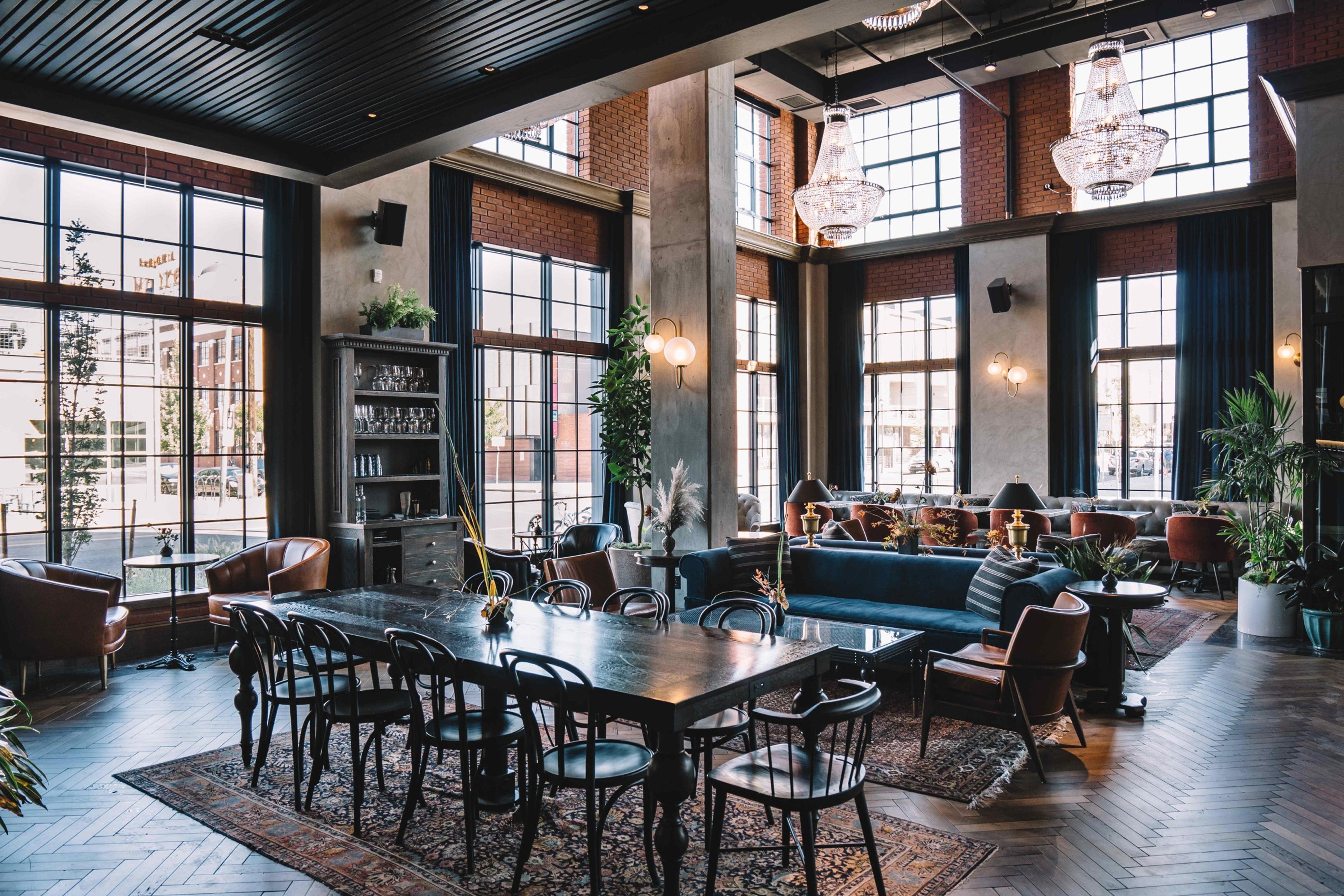 A high ceilinged lounge with tall arched windows, two chandeliers, a long wooden dining table and chairs, and a blue sofa against a brick wall.