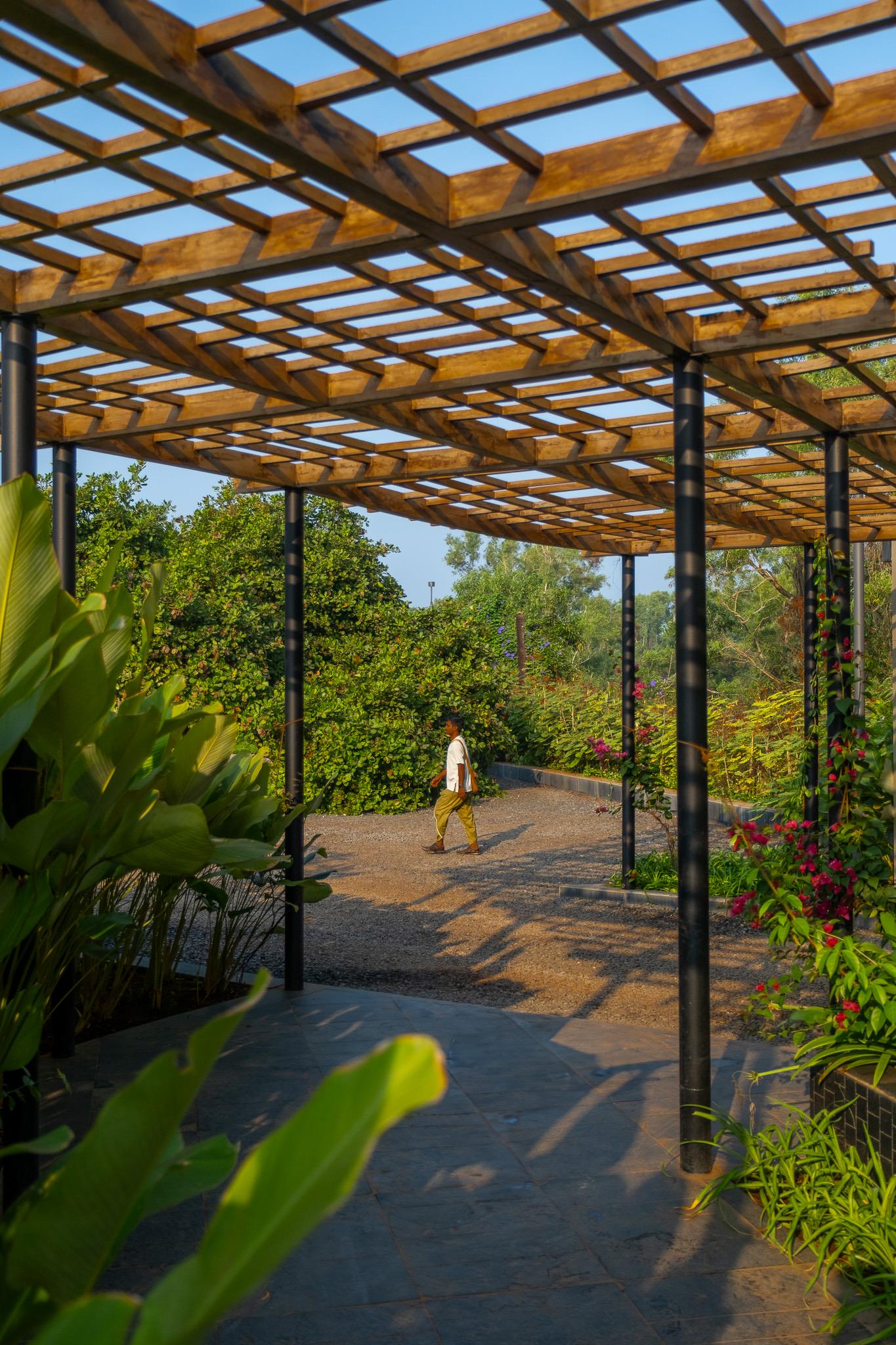 A wooden lattice pergola over a stone path, with a person walking beneath it towards flowering shrubs and trees.
