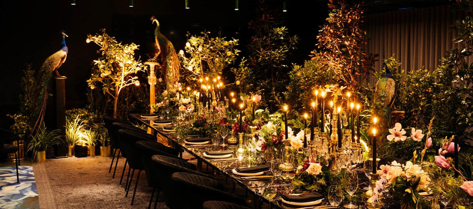 a long candlelit dining table set among potted trees and taxidermy peacocks, laid with glassware and flowers