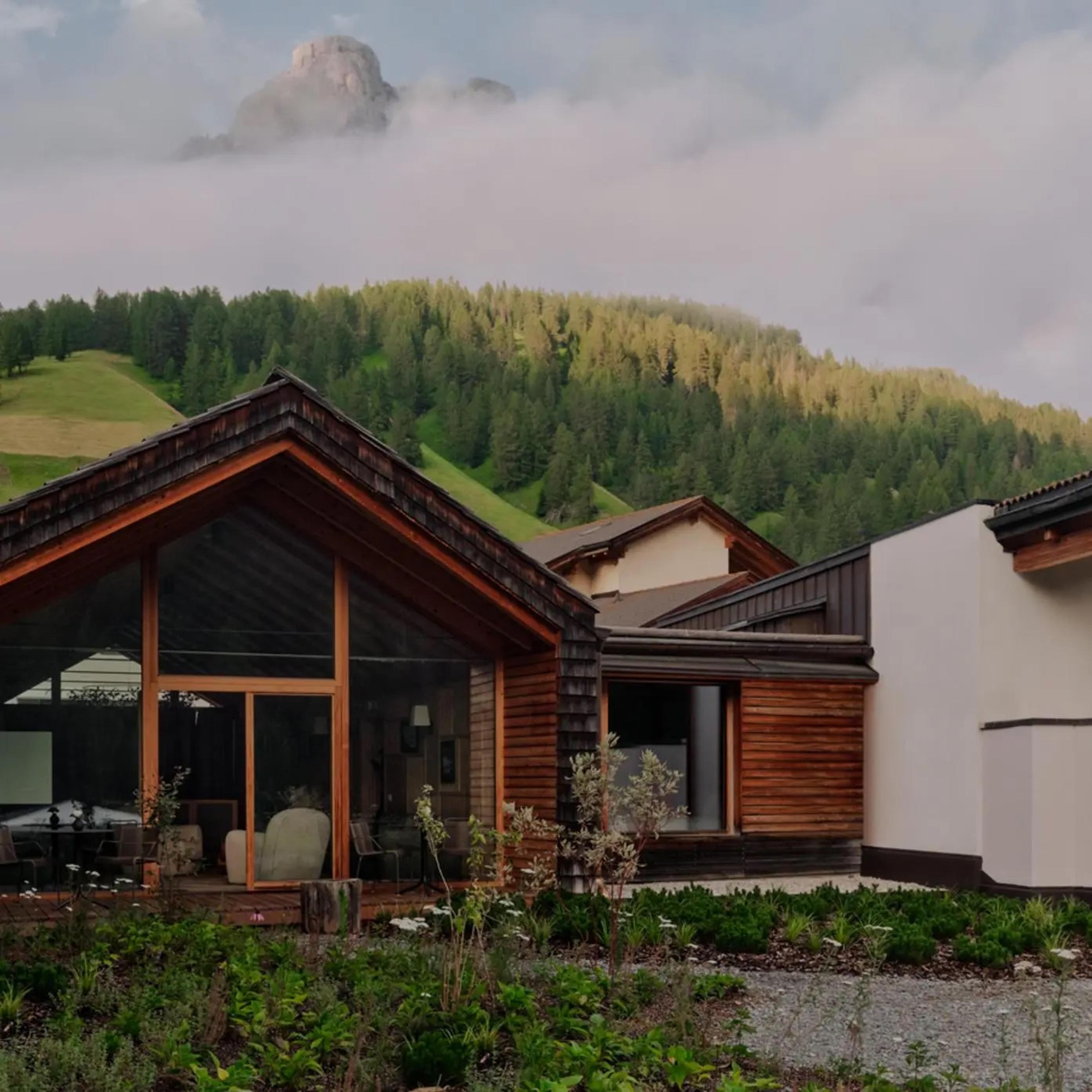 a small timber and stone chalet with a garden of wildflowers in front, a forested hillside and cloud wreathed mountain peak behind