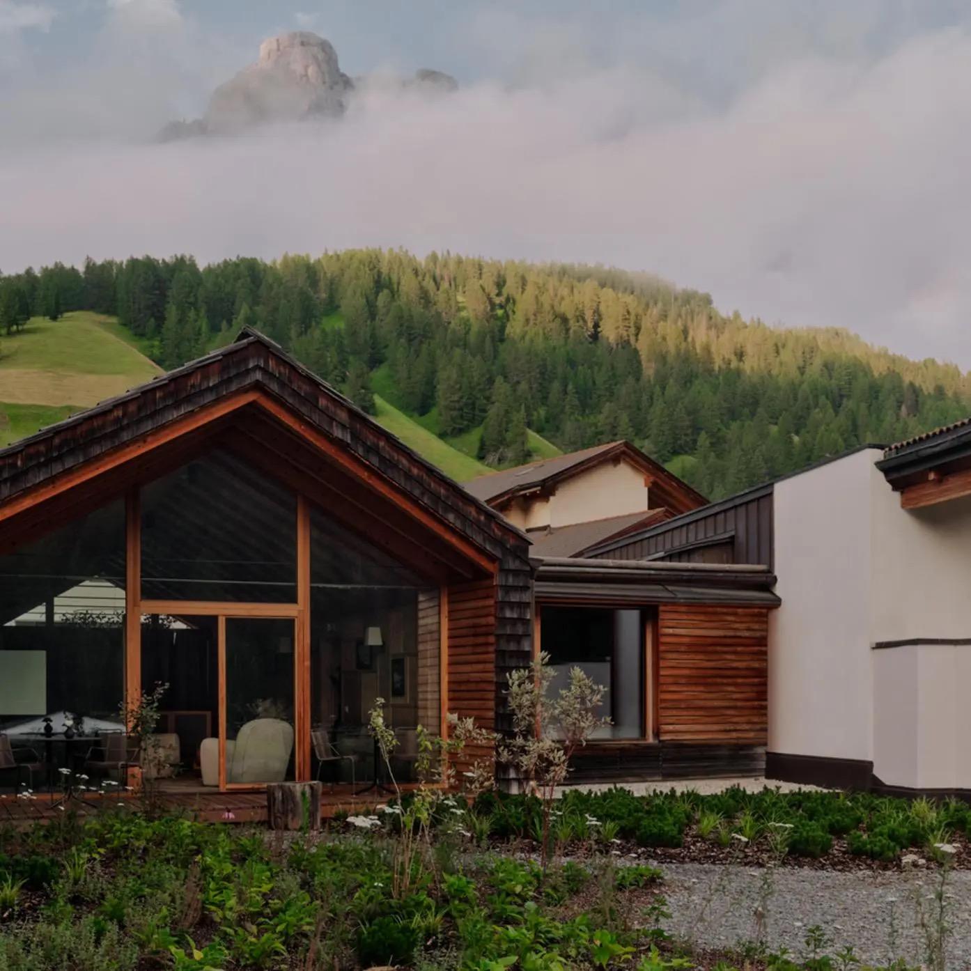 a small timber and stone chalet with a garden of wildflowers in front, a forested hillside and cloud wreathed mountain peak behind