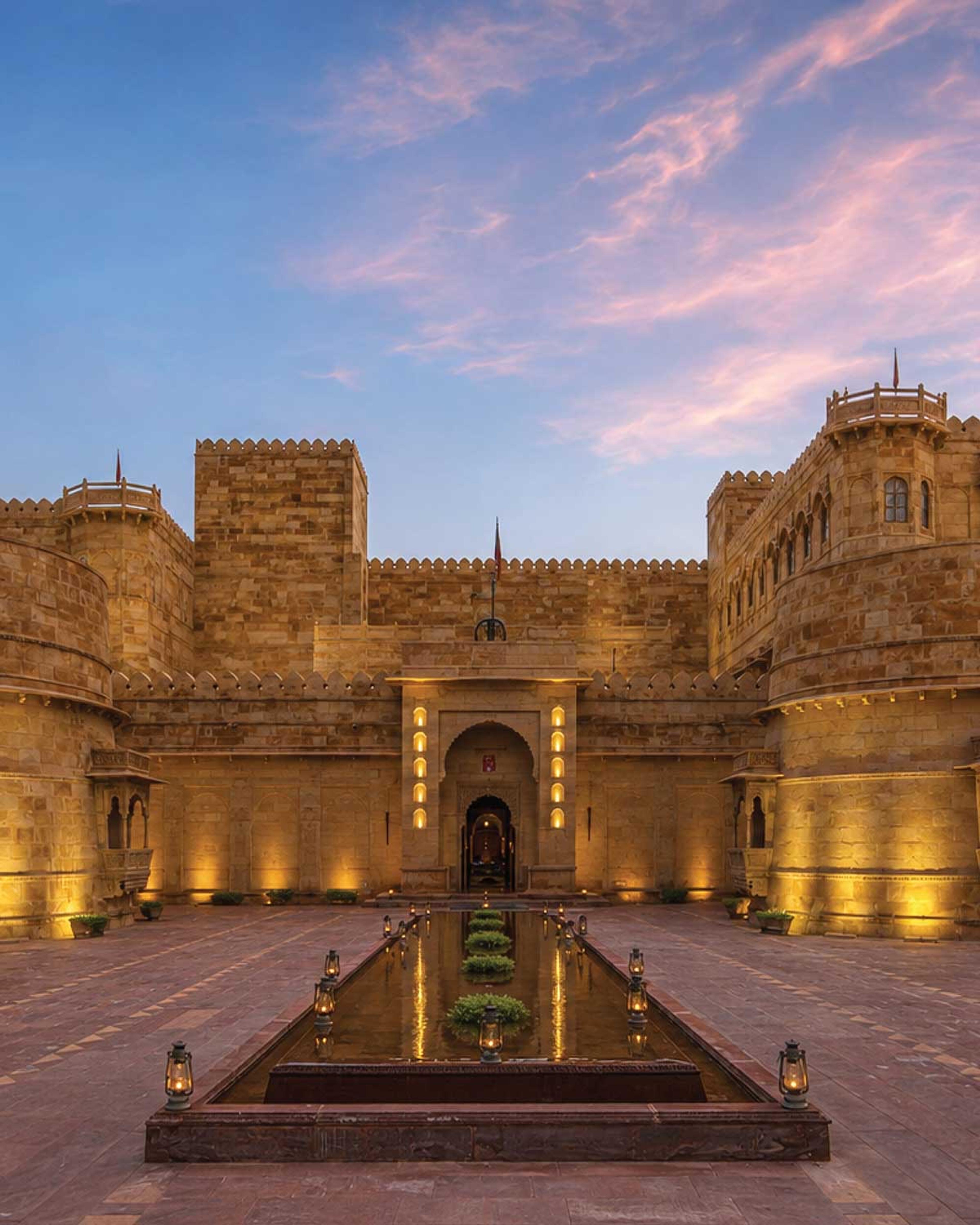 the sandstone palace lit at dusk, its turrets and battlements against a pink and blue sky, with a long reflecting pool and lanterns leading to the entrance
