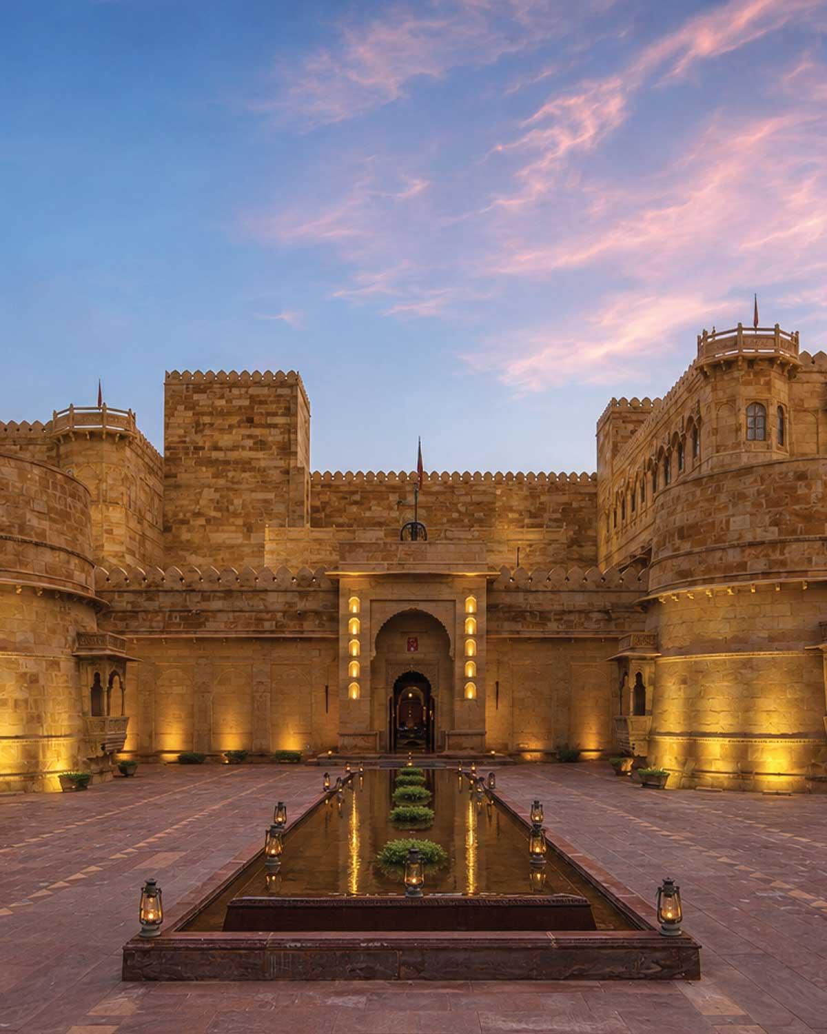 the sandstone palace lit at dusk, its turrets and battlements against a pink and blue sky, with a long reflecting pool and lanterns leading to the entrance
