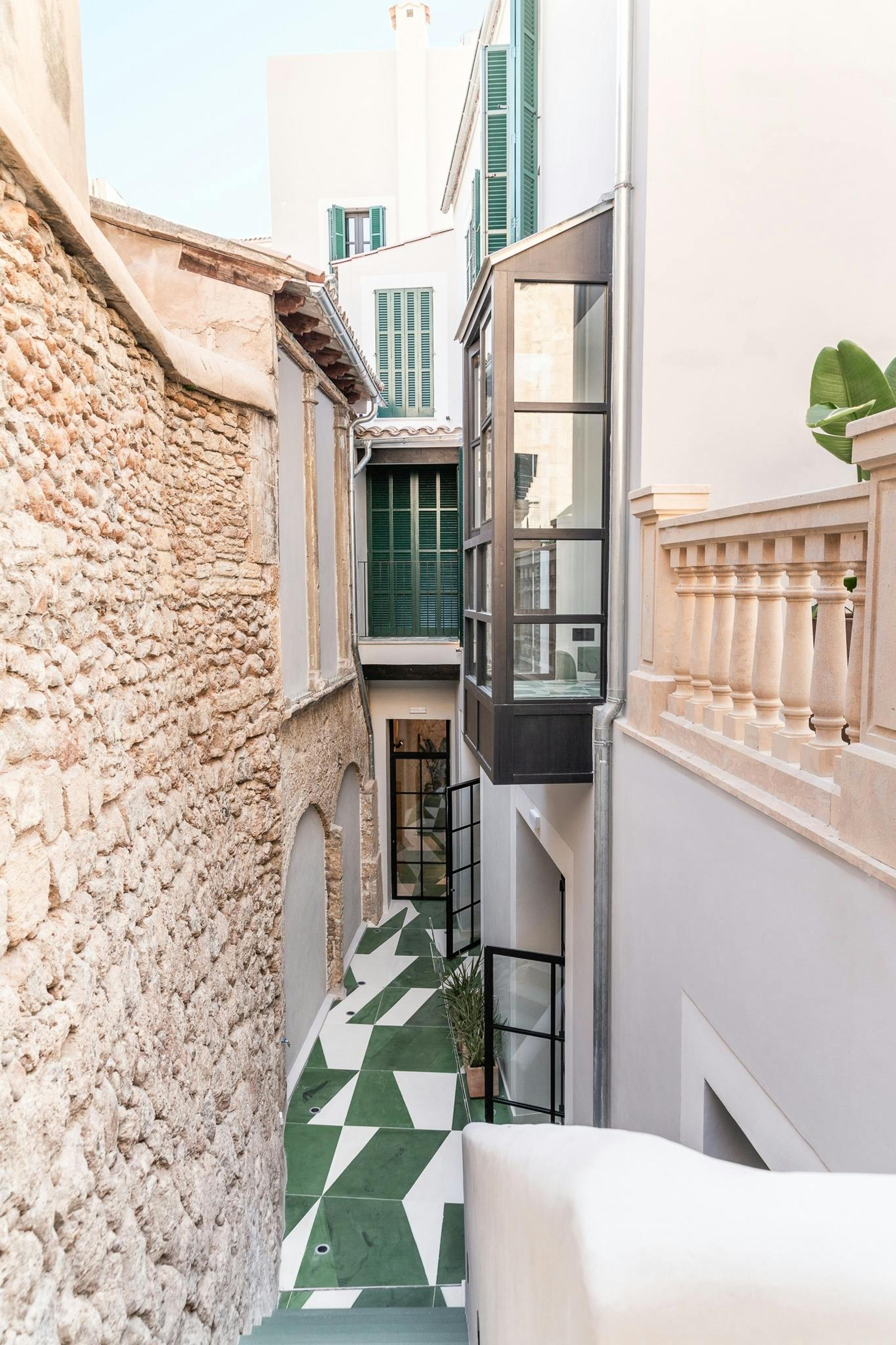 A narrow lane between a stone wall and a white building with green shutters, its floor laid in a green and white geometric tile pattern leading toward the hotel.