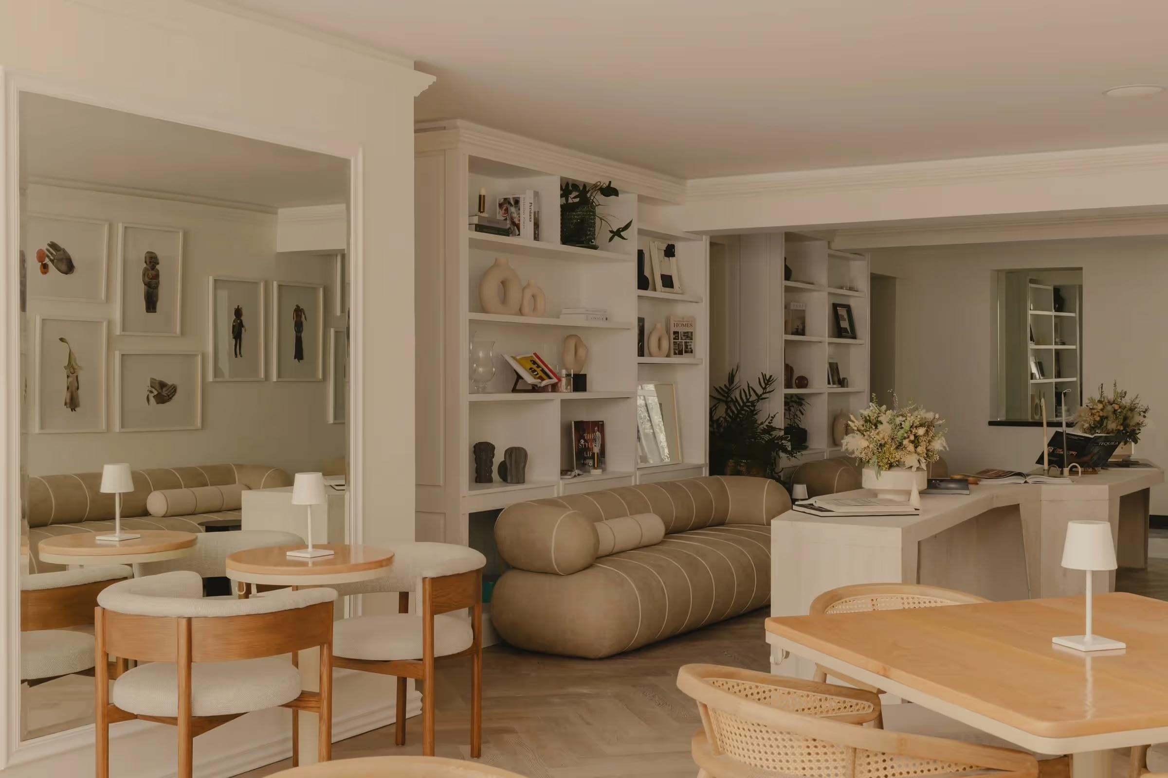 A hotel lounge with built-in shelving holding small sculptures and books, a low sofa, round tables and wooden dining chairs, reflected in a large mirror.