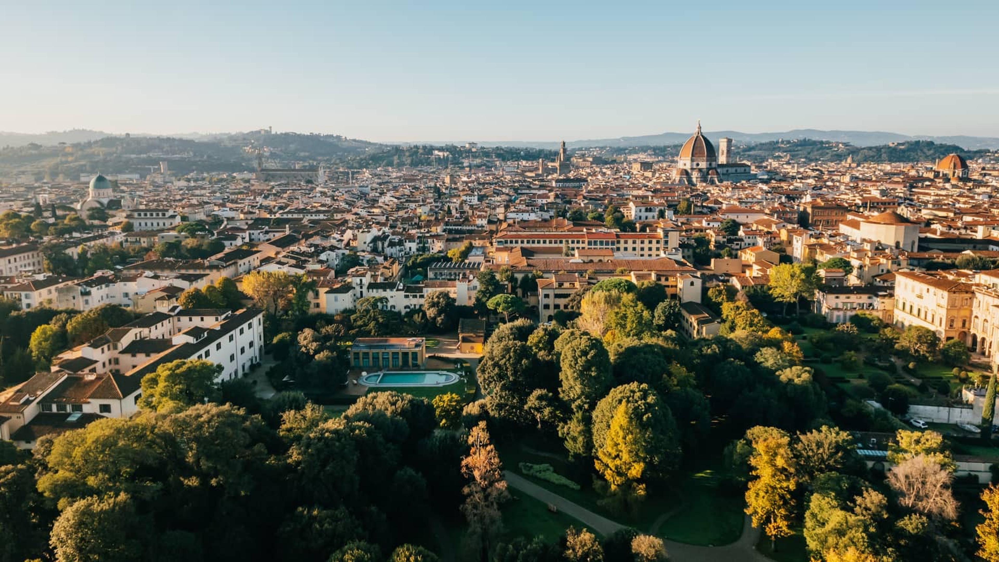 An aerial view over Florence at golden hour, showing a private garden and rectangular pool among trees with the city's rooftops and the cathedral dome beyond.