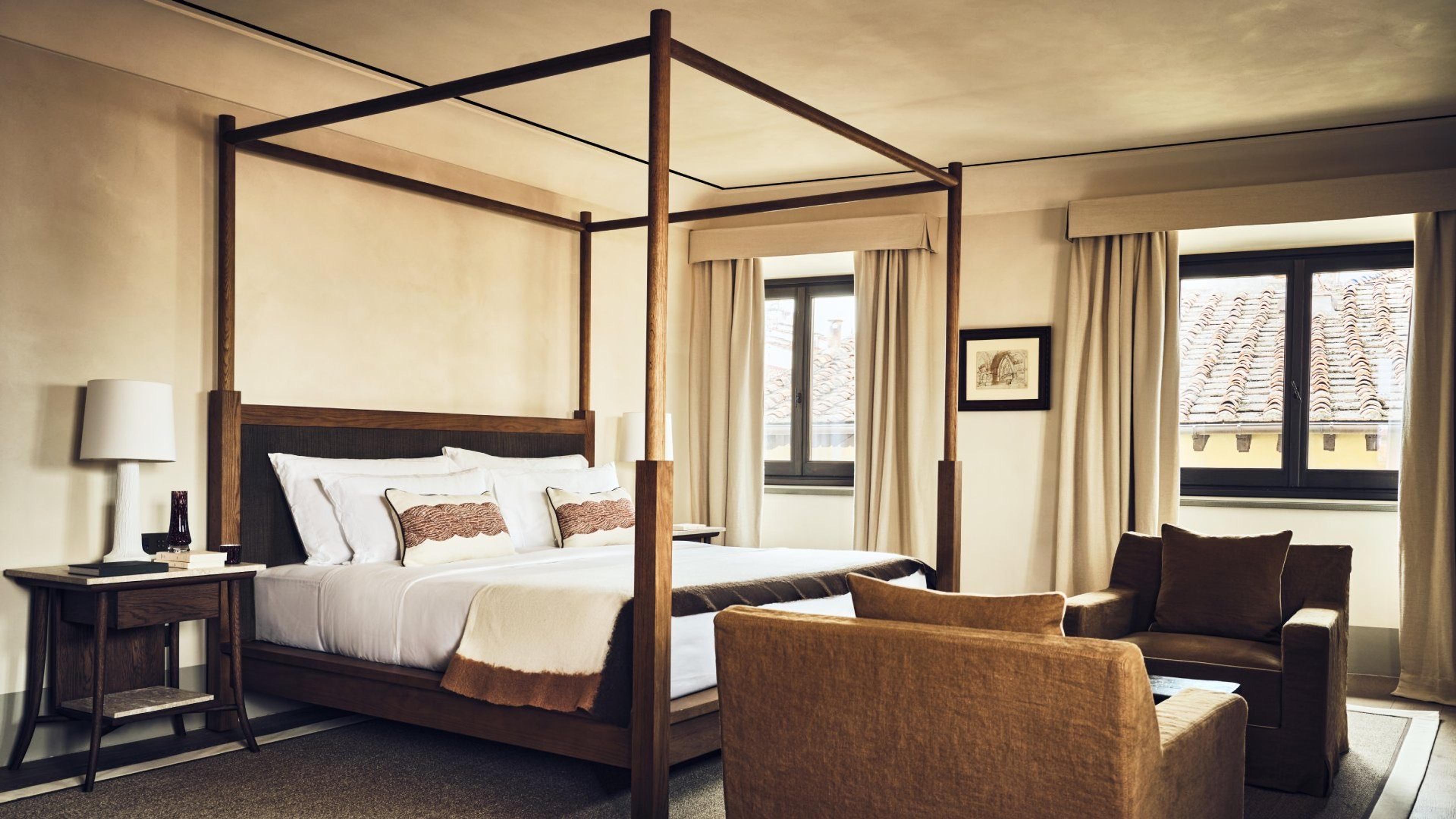 A four-poster bed and two brown armchairs in a high-ceilinged room, with two open windows looking out over terracotta rooftops.