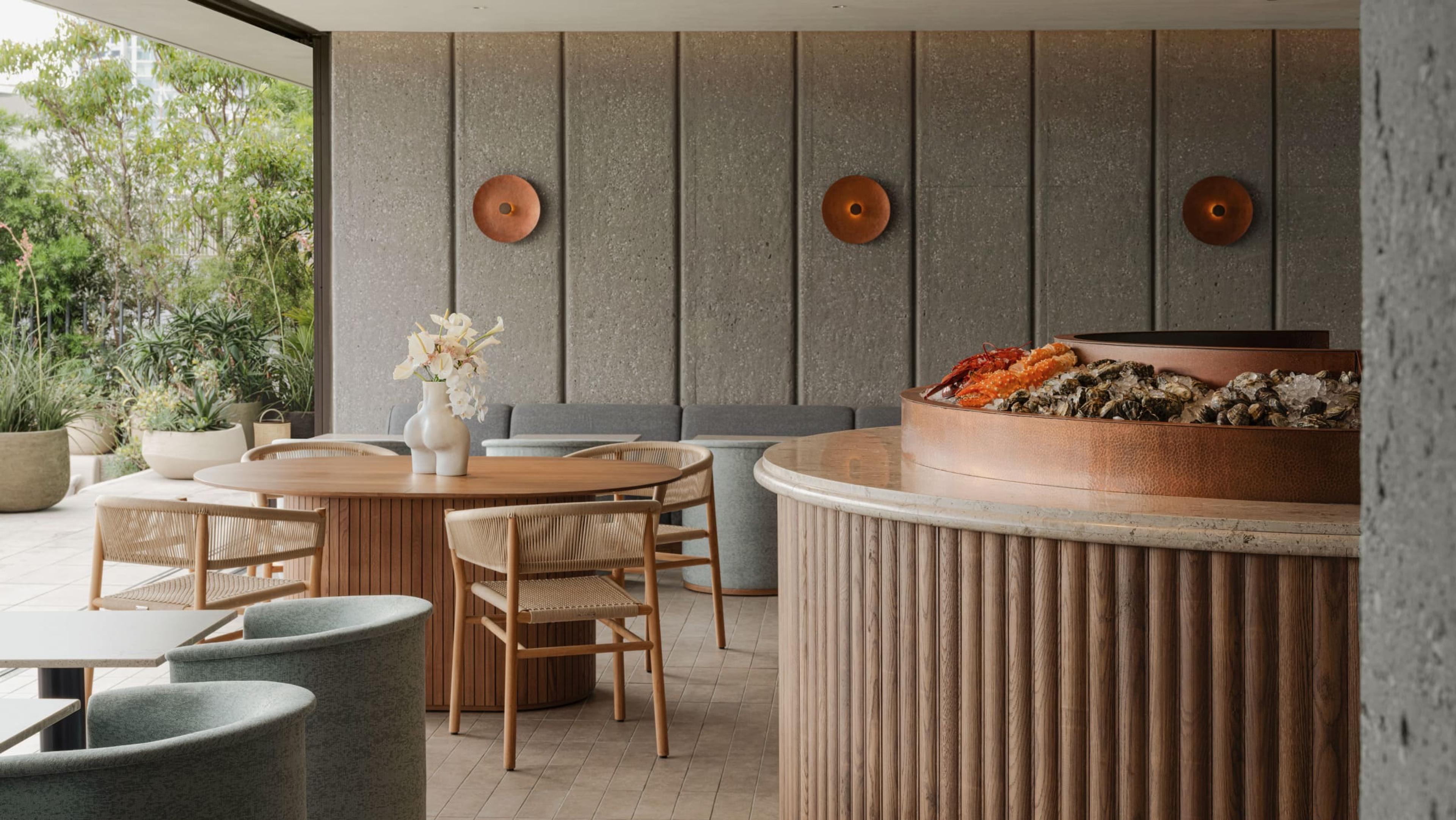 a round copper raw bar heaped with oysters and shellfish on ice, beside a wood dining table set with a vase of white flowers