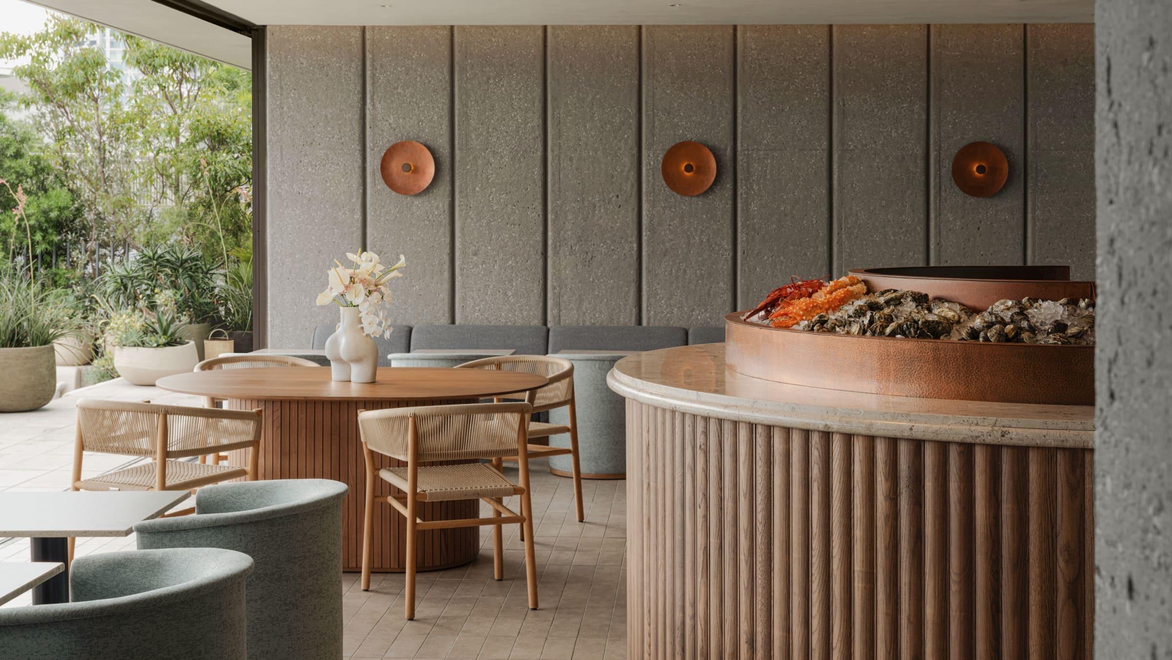 a round copper raw bar heaped with oysters and shellfish on ice, beside a wood dining table set with a vase of white flowers