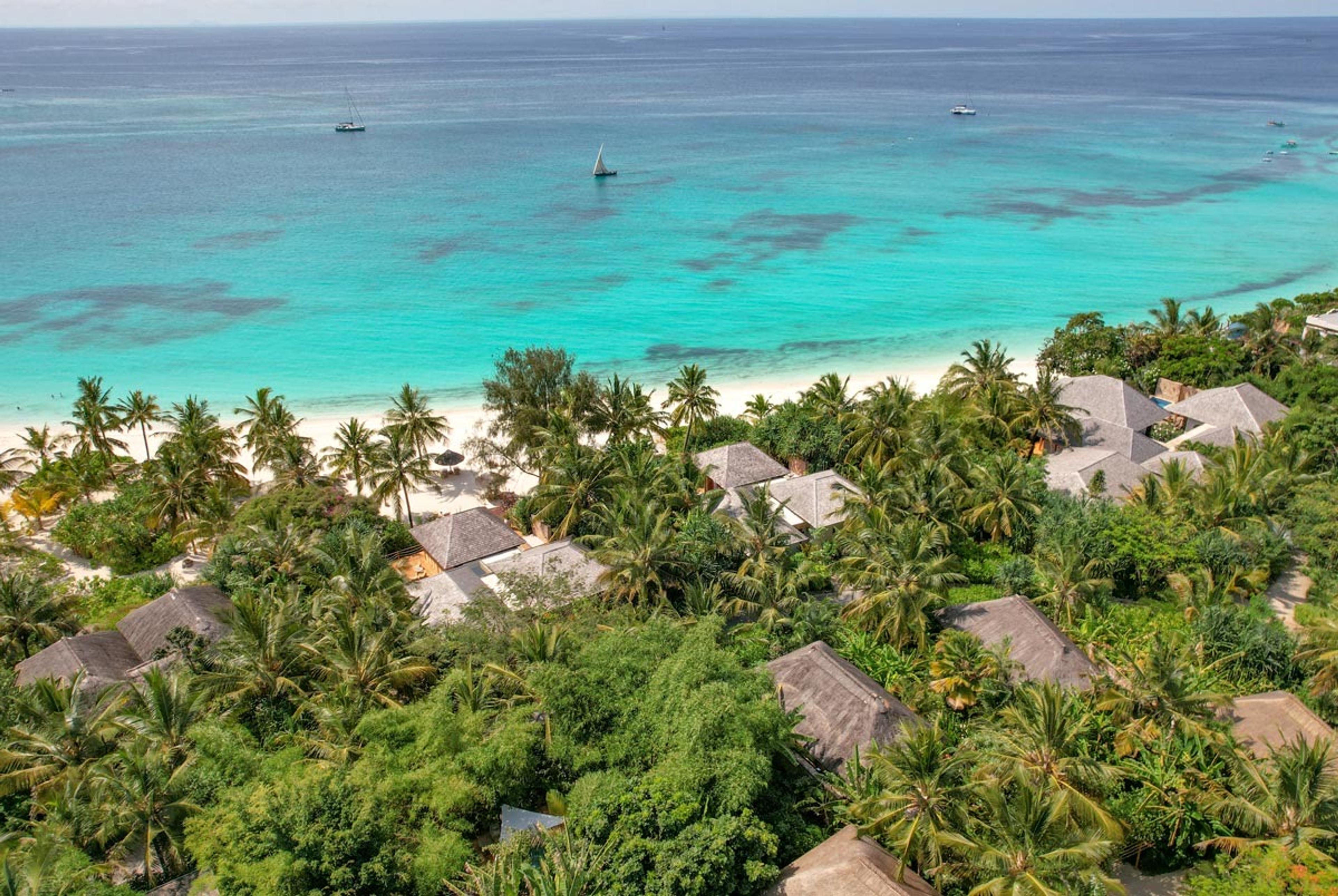 an aerial view of a resort's grey thatched roofs set among palm trees, with a white sand beach and two sailing dhows on the turquoise sea beyond