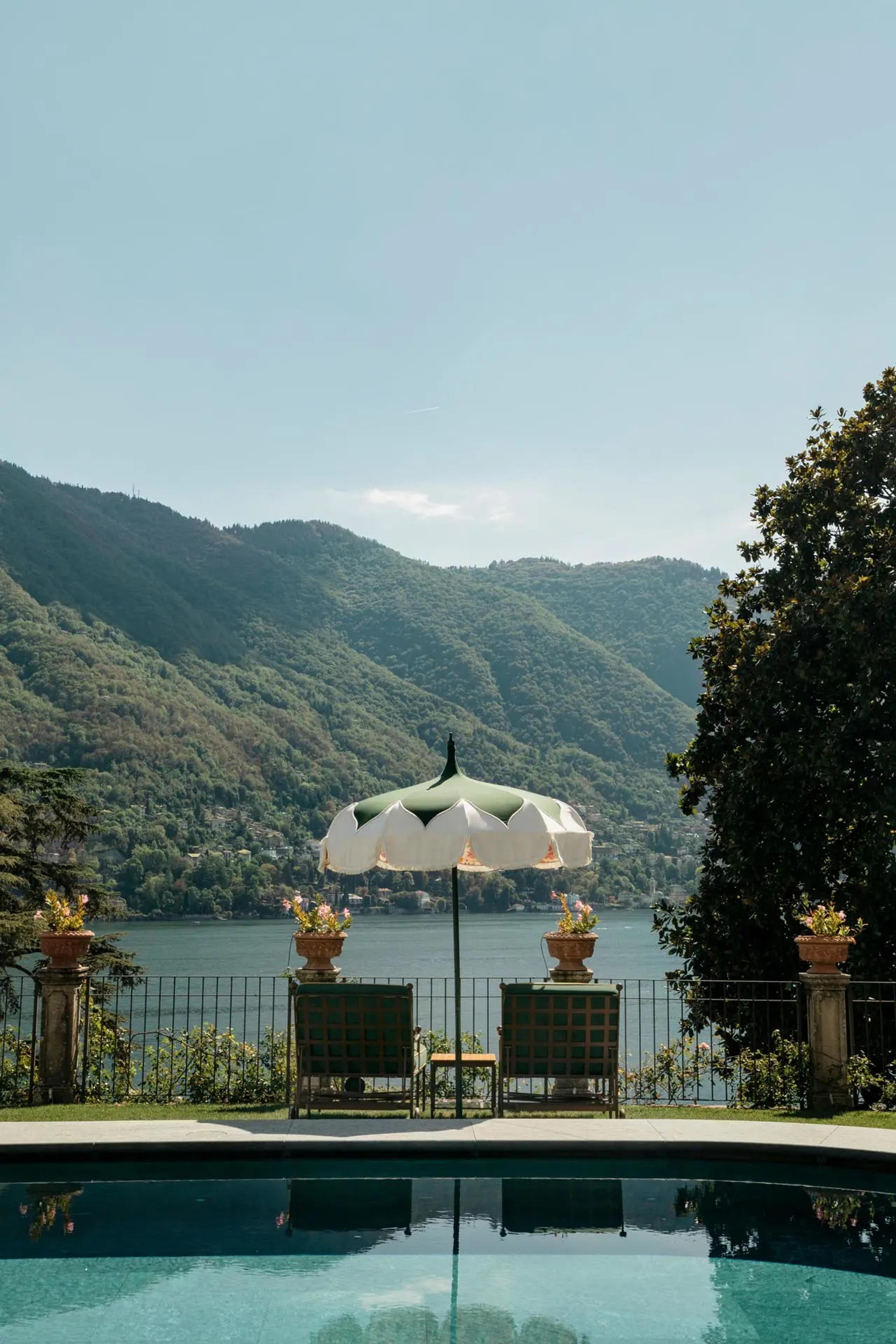 A poolside terrace with a scalloped umbrella and two chairs behind a railing, looking across a lake to wooded mountains.