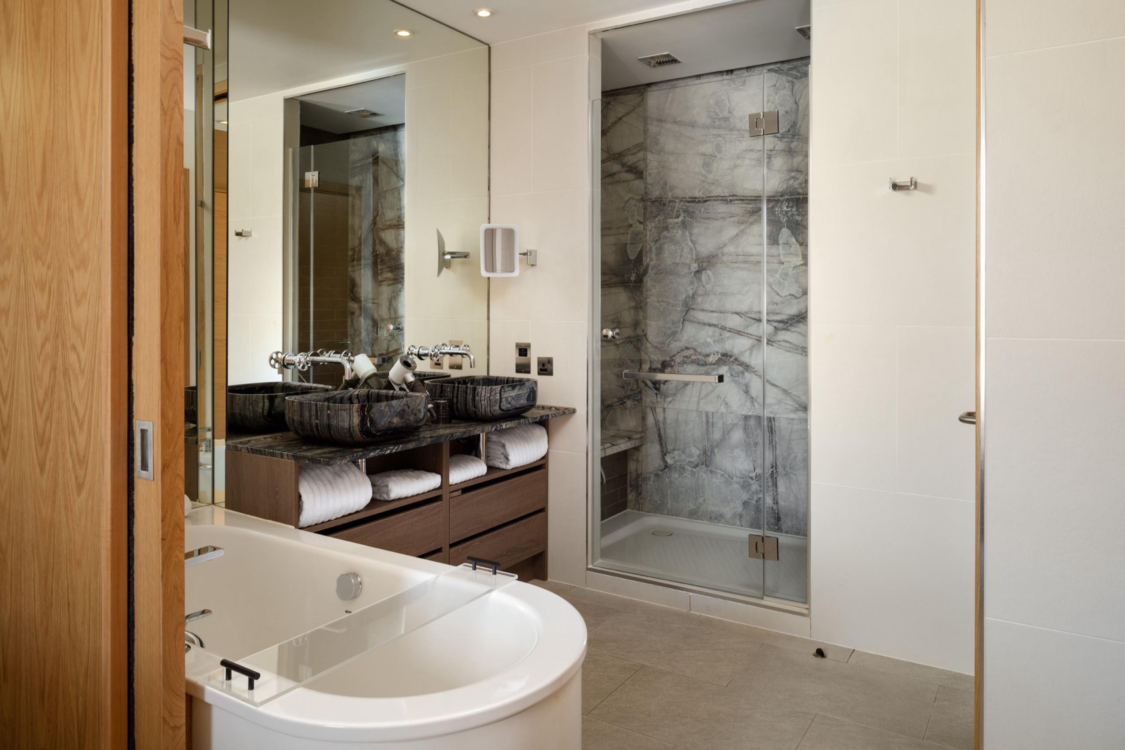A freestanding white bathtub beside two marble sinks under a large mirror, with a walk-in double shower alongside.