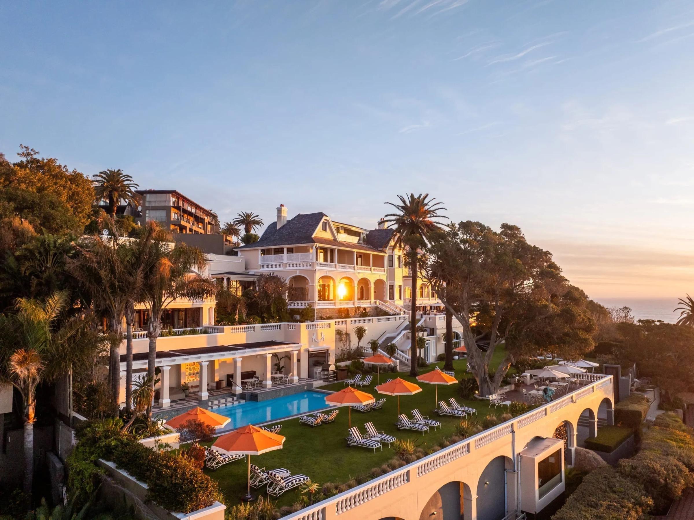 An aerial view at sunset of a long cream building with arched colonnades and a pool with orange parasols, set on a terraced lawn above trees, with the sea at the far edge.