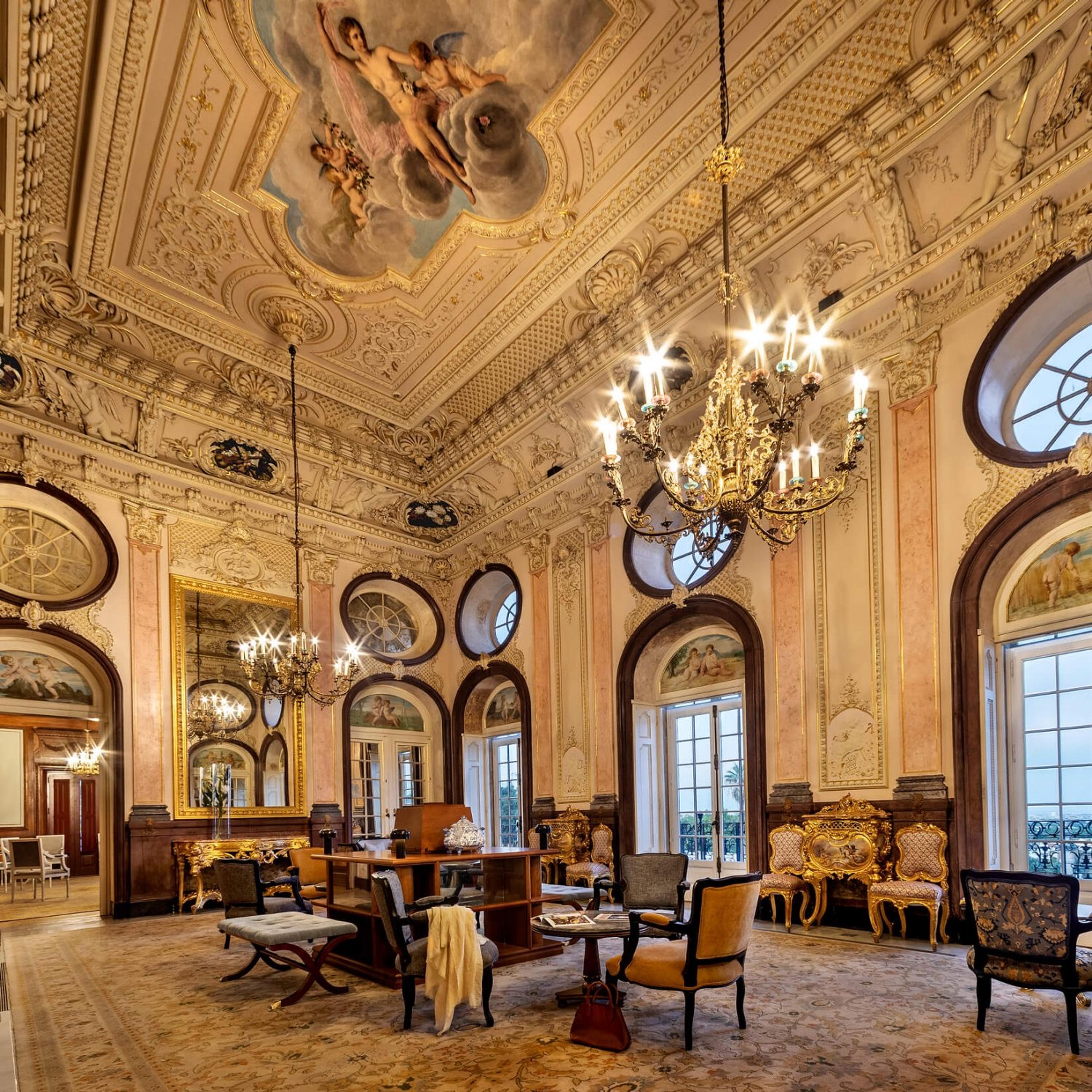 A high lounge with a painted ceiling of cherubs, gilt chandeliers, a large gilt-framed mirror, and armchairs on a patterned rug.