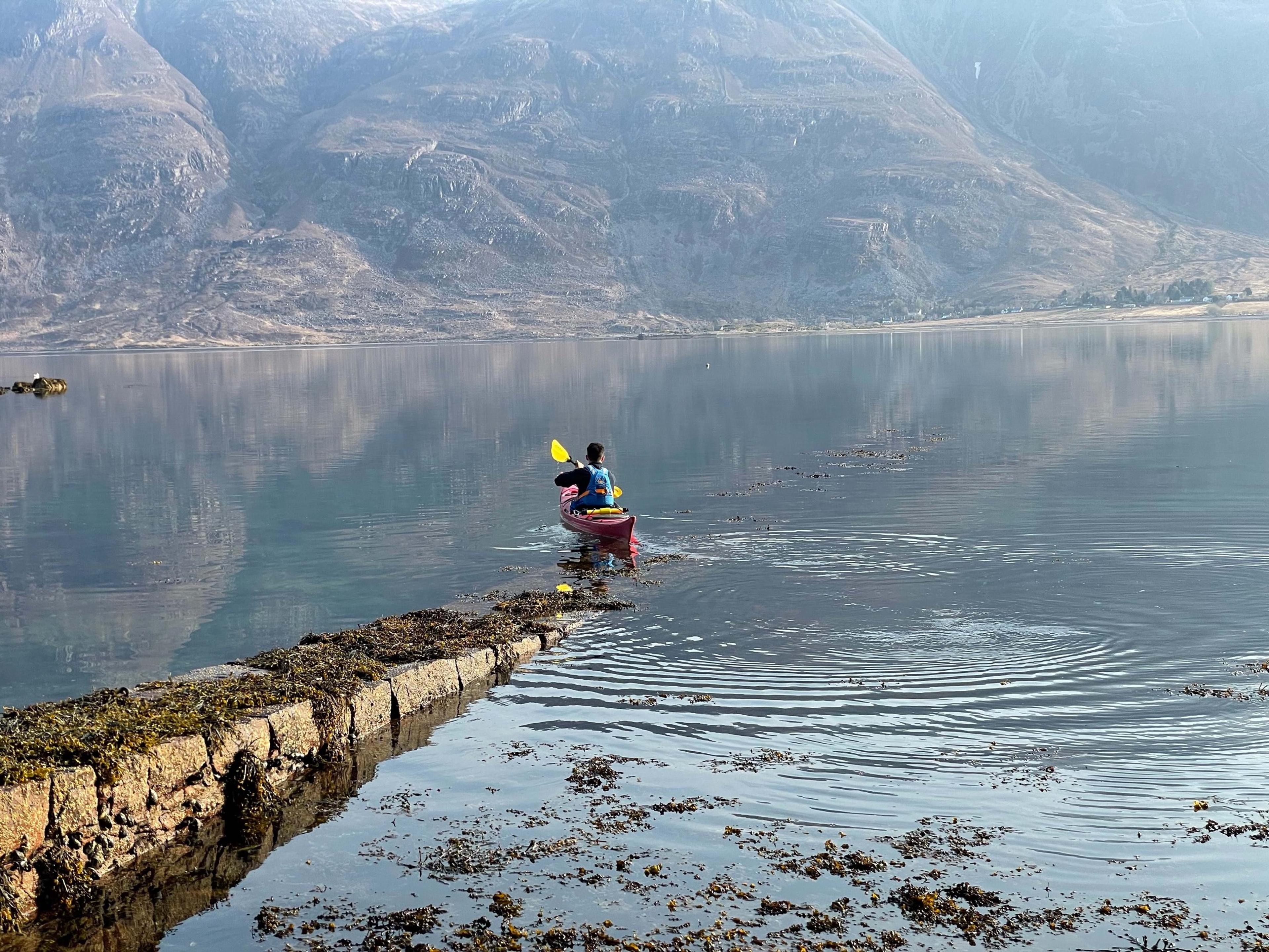 A person kayaking on a still loch at dawn, paddling away from a stone jetty with mountains reflected in the water.