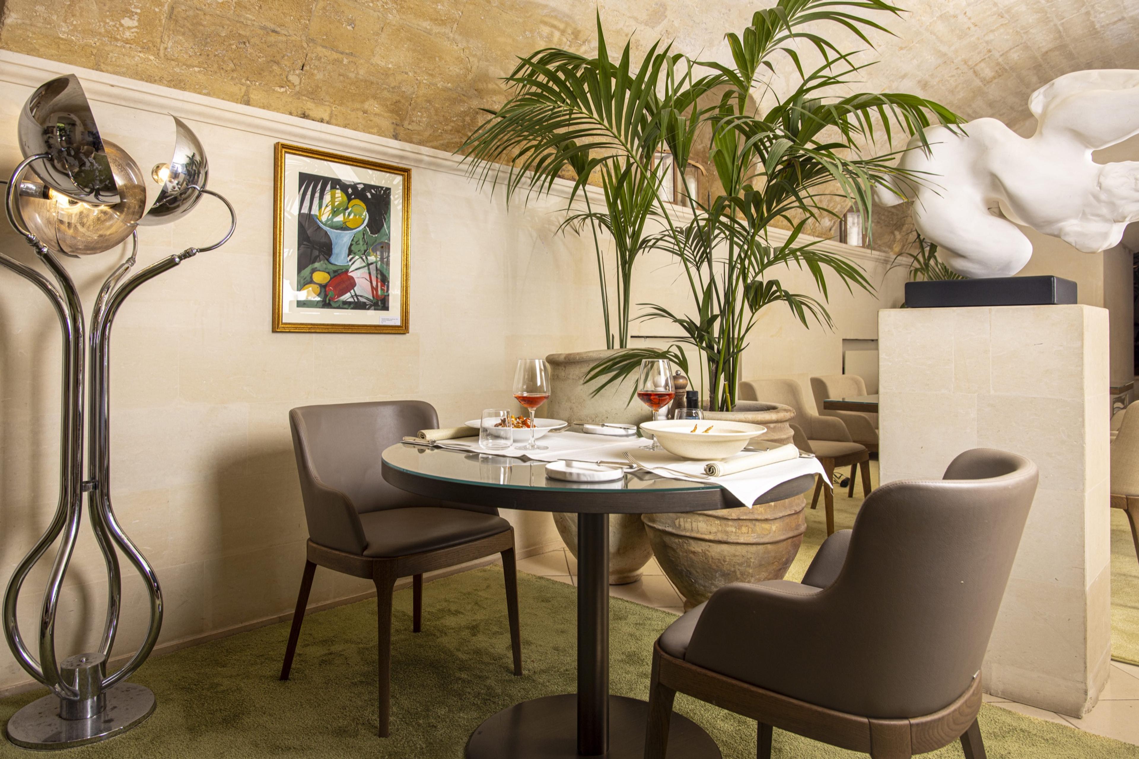 A small dining alcove with a vaulted stone ceiling, a round table set with wine glasses, a potted palm and a nude statue nearby.