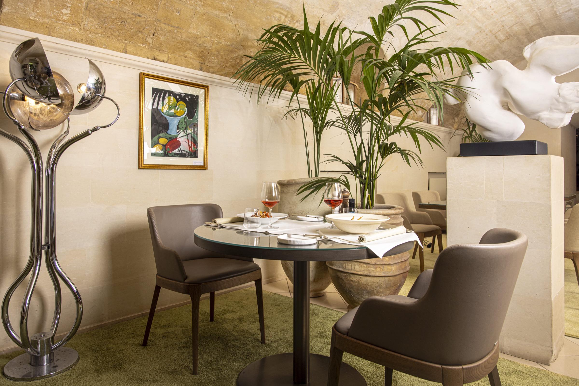 A small dining alcove with a vaulted stone ceiling, a round table set with wine glasses, a potted palm and a nude statue nearby.